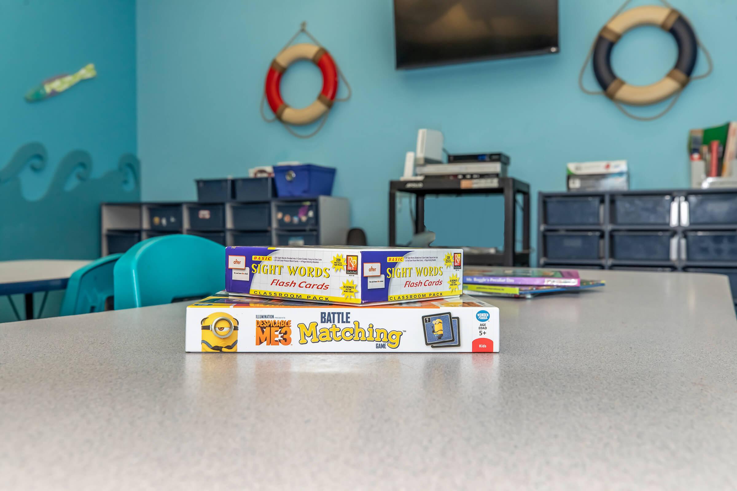 Flashcards for sight words and a matching game are stacked on a table in a brightly colored classroom. The background features a TV mounted on the wall and storage units, with playful décor such as lifebuoys, creating a cheerful learning environment.