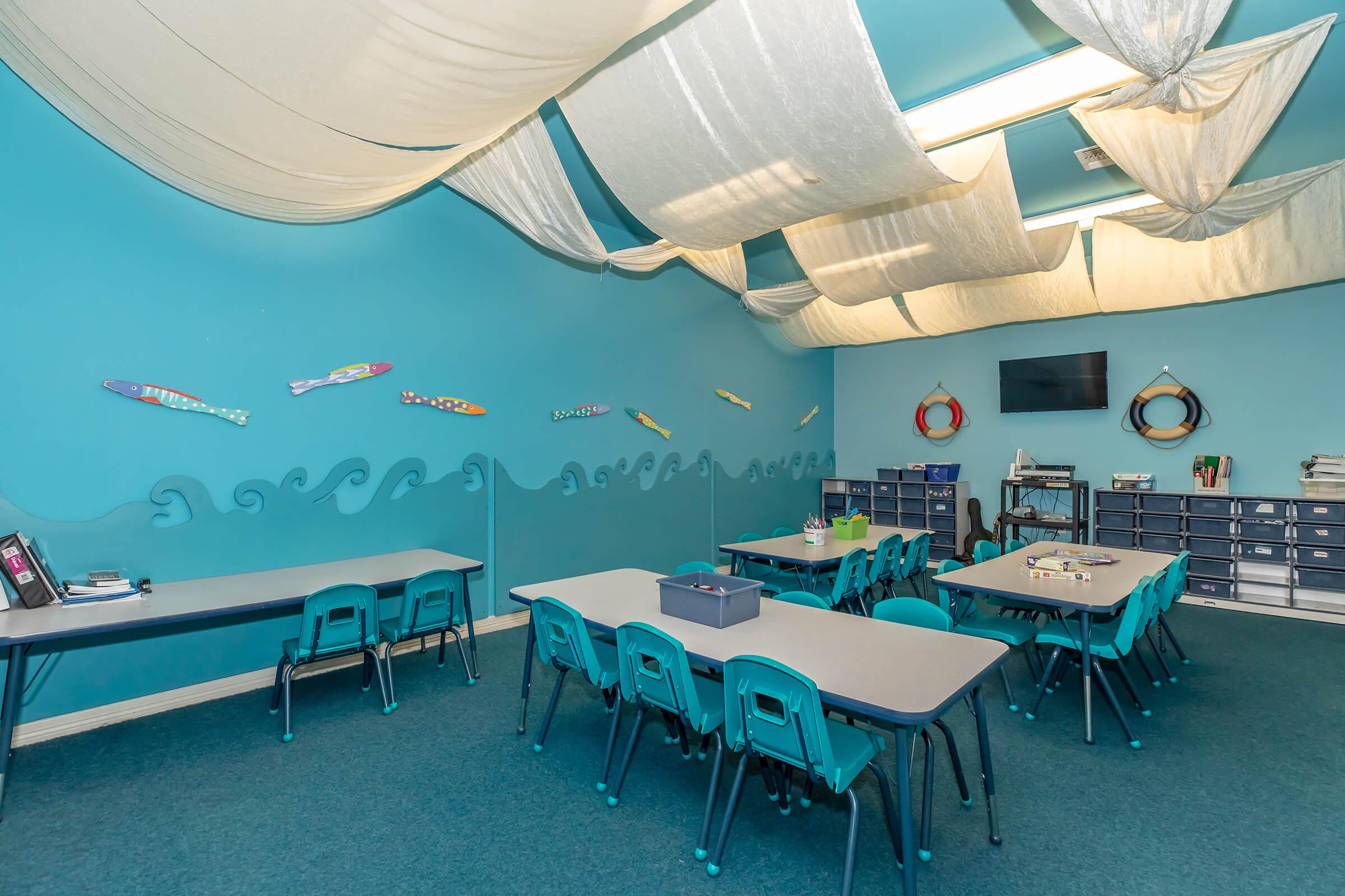 A bright classroom with turquoise walls and a ceiling decorated with draped fabric. Several tables and teal chairs are arranged for students. The walls feature colorful fish decorations and a wave pattern. There is a TV and a storage unit with drawers in the room, creating an inviting learning environment.