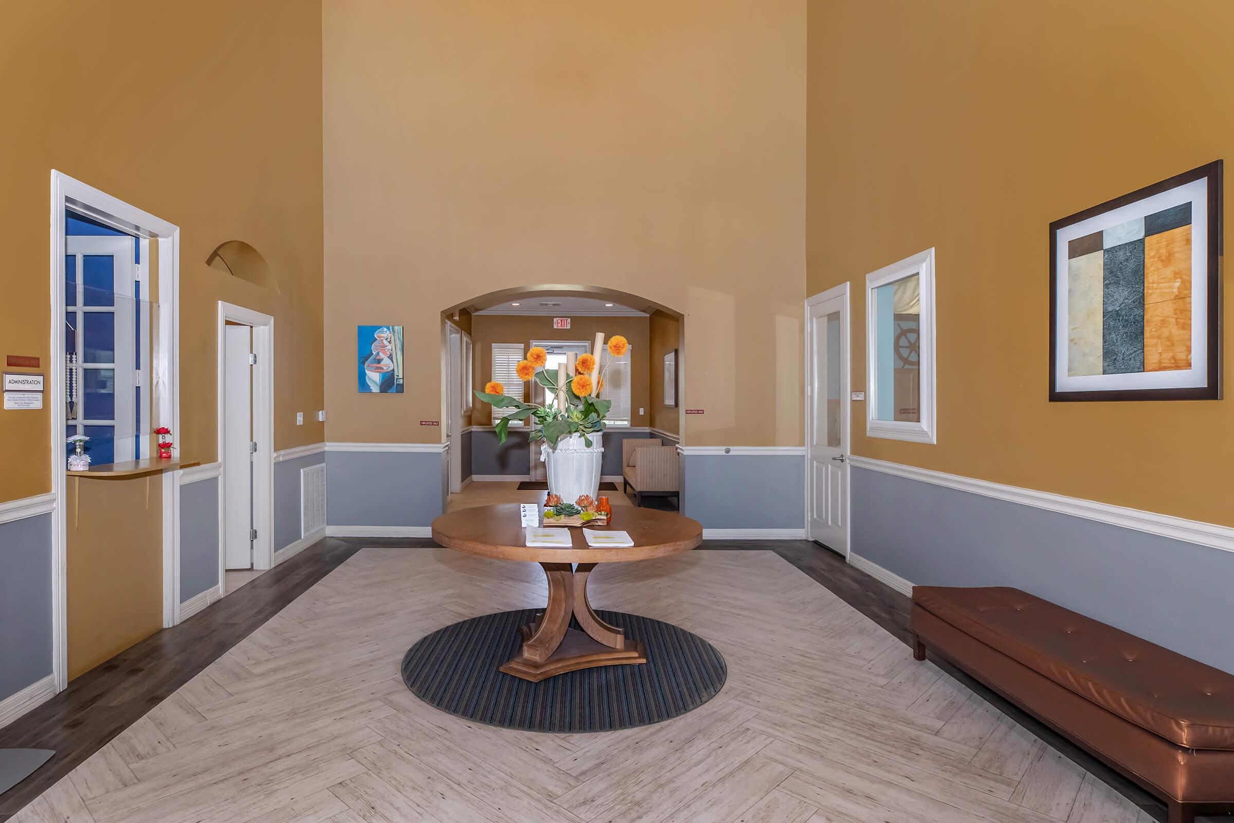 A bright and spacious reception area featuring a round wooden table with a floral arrangement at its center. The walls are painted in warm tones, and there are decorative elements including artwork and a brown leather bench. Doors lead to other rooms, enhancing the welcoming atmosphere.