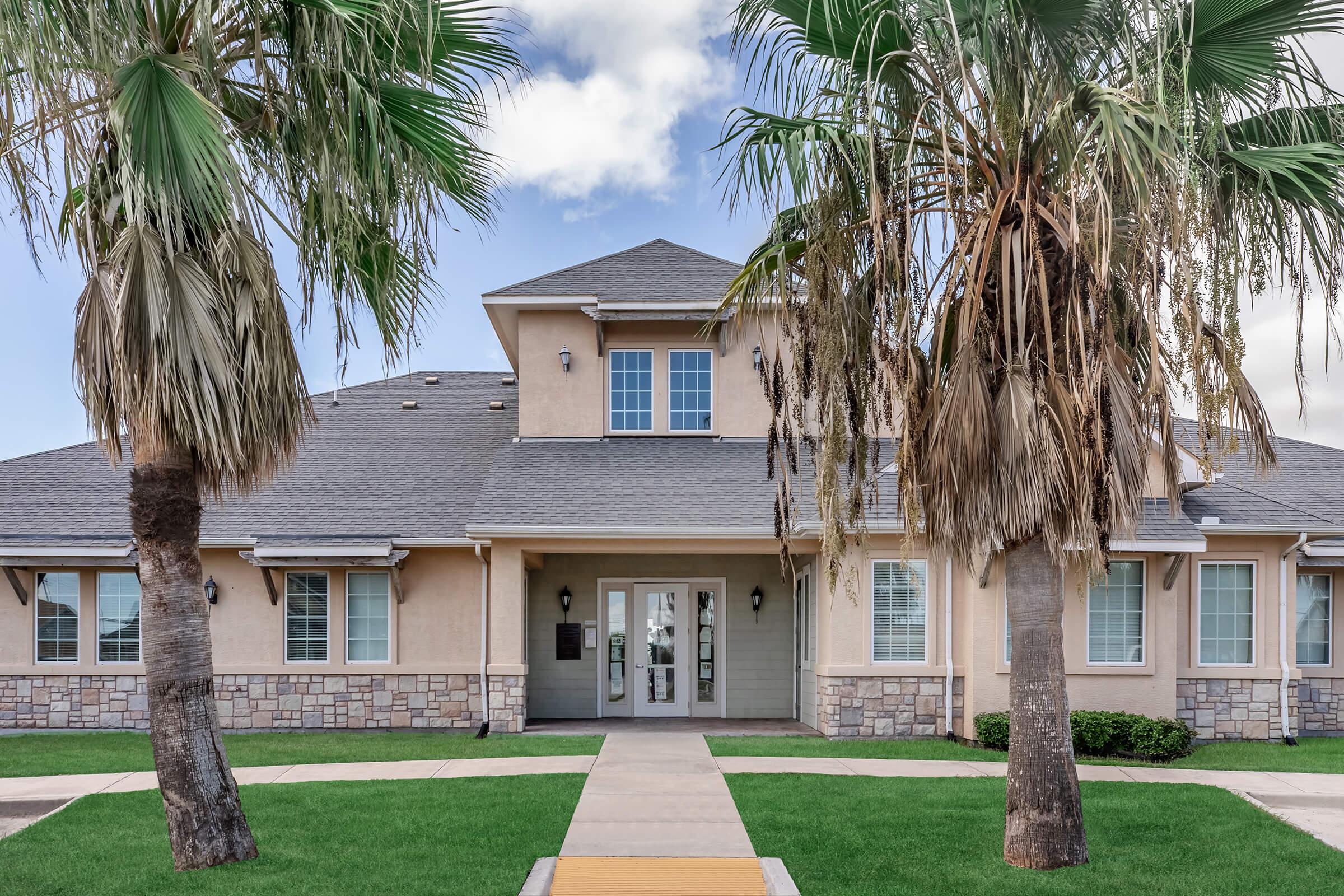 A well-maintained building with a light-colored facade, surrounded by palm trees. The entrance features large windows and a welcoming porch. A paved pathway leads to the door, with well-manicured green grass on either side, set against a partly cloudy sky.
