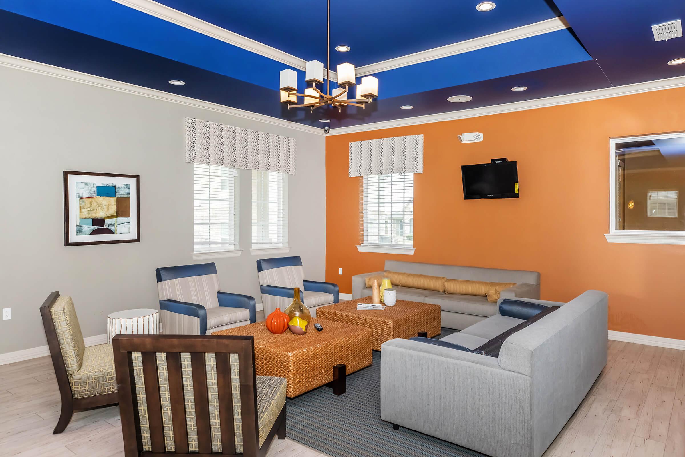 A modern living room with a mix of blue and orange walls, featuring a gray sofa, blue chairs, and a woven coffee table. Decor includes a framed artwork, a small pumpkin, and decorative pillows. There are large windows with blinds, a flat-screen TV mounted on the wall, and pendant lighting hanging from the ceiling.