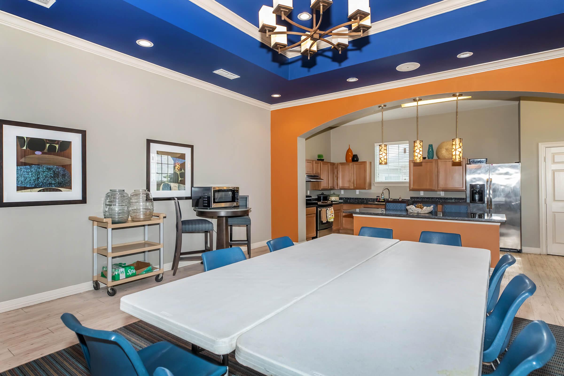 A modern communal kitchen and dining area featuring a large white table surrounded by blue chairs. The walls are painted in neutral colors with an accent orange arch. The kitchen has wooden cabinets, a microwave, and stainless steel appliances, creating a welcoming atmosphere.