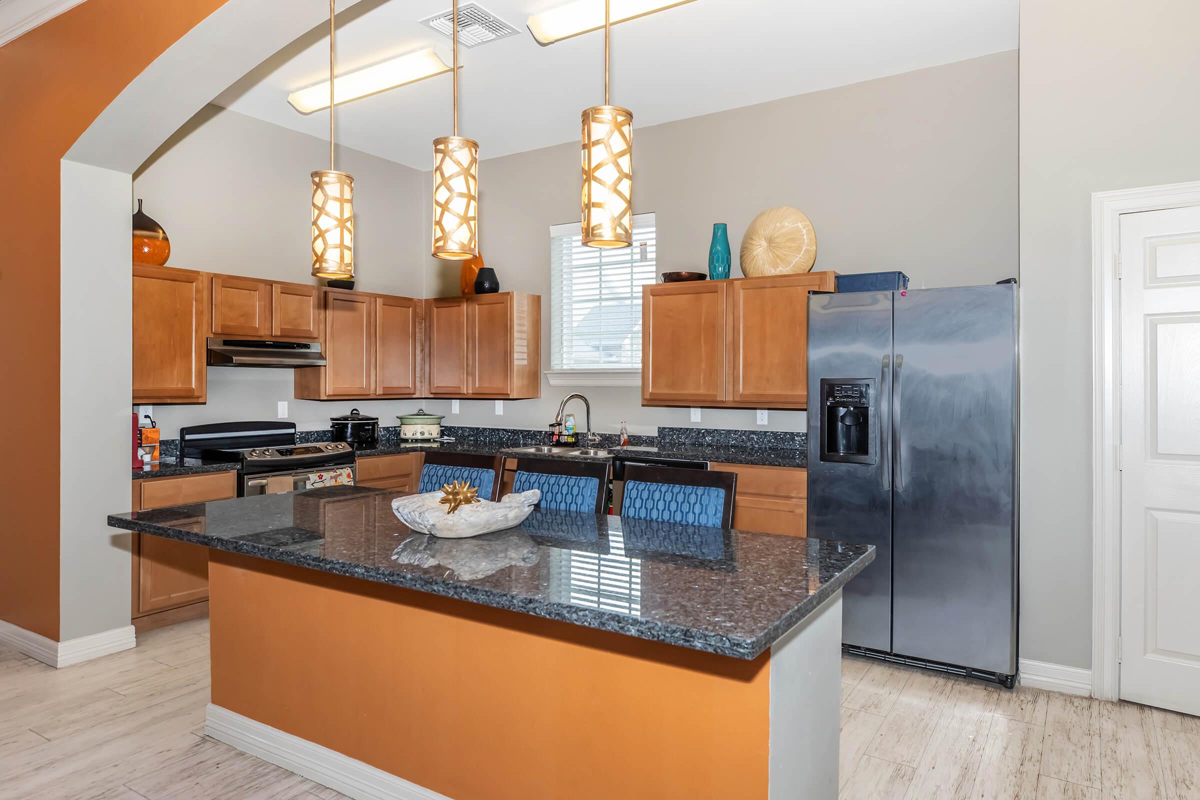 Modern kitchen featuring wooden cabinets, granite countertops, and stainless steel appliances. Pendant lights hang above an island with blue bar stools. The wall has an orange color and decorative items are placed on the countertops. Natural light enters through a window, illuminating the space.
