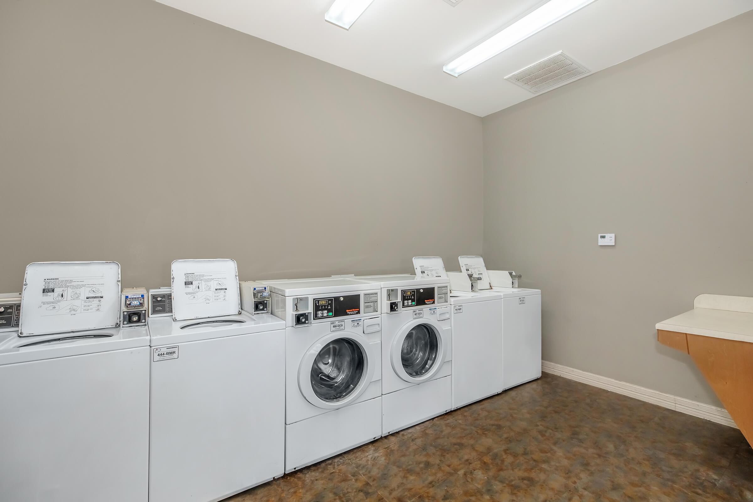 A clean and well-lit laundry room featuring three white washing machines and a dryer. The machines are lined up against a neutral-colored wall, and there is a folding table on the right. The space appears organized and functional, suitable for doing laundry.
