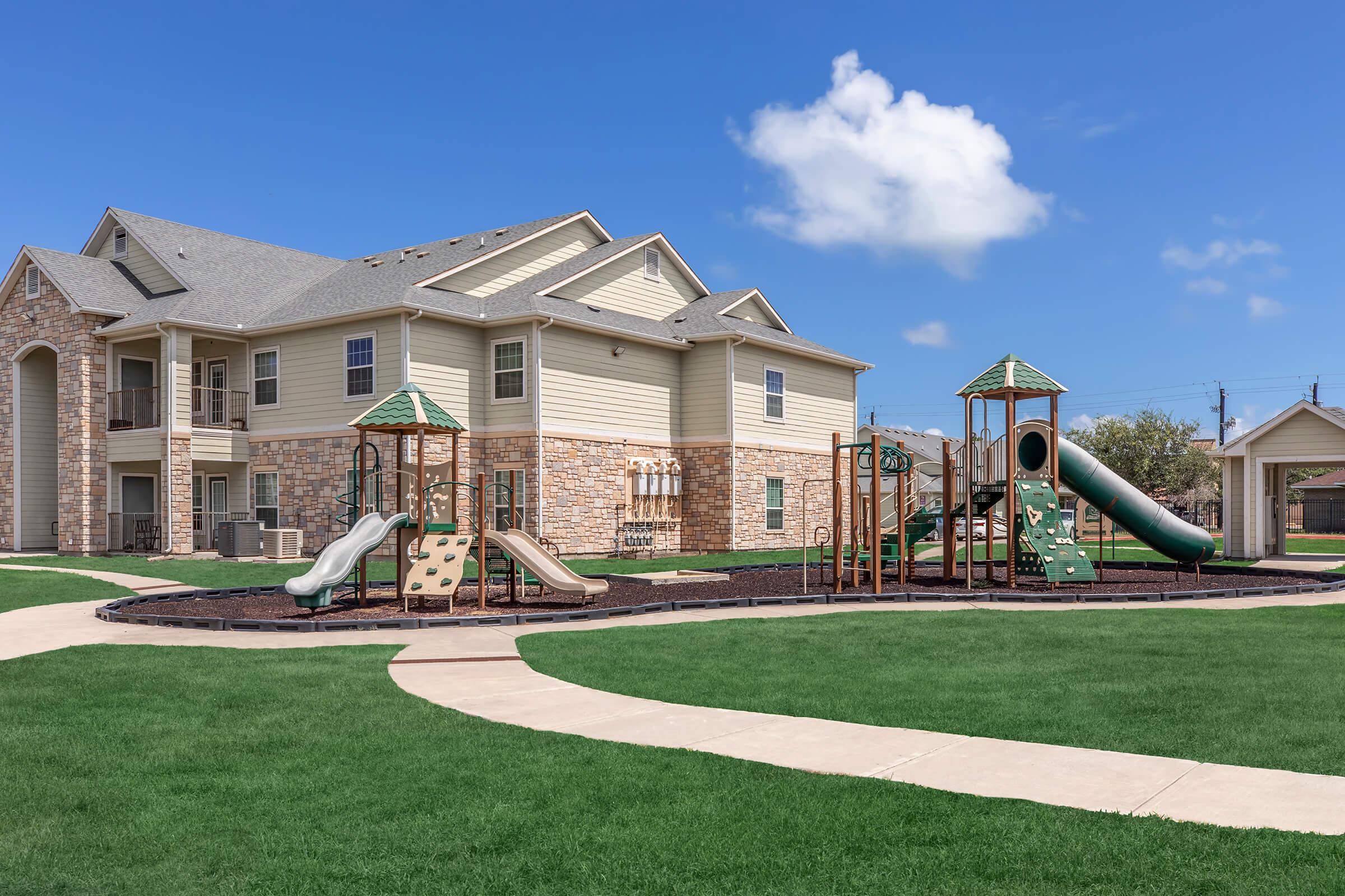 Playground area with slides and climbing structures surrounded by green grass, with a residential building in the background. The sky is clear and blue with a few clouds, creating a bright and inviting atmosphere. Pathways wind through the landscape, providing access to the play equipment.