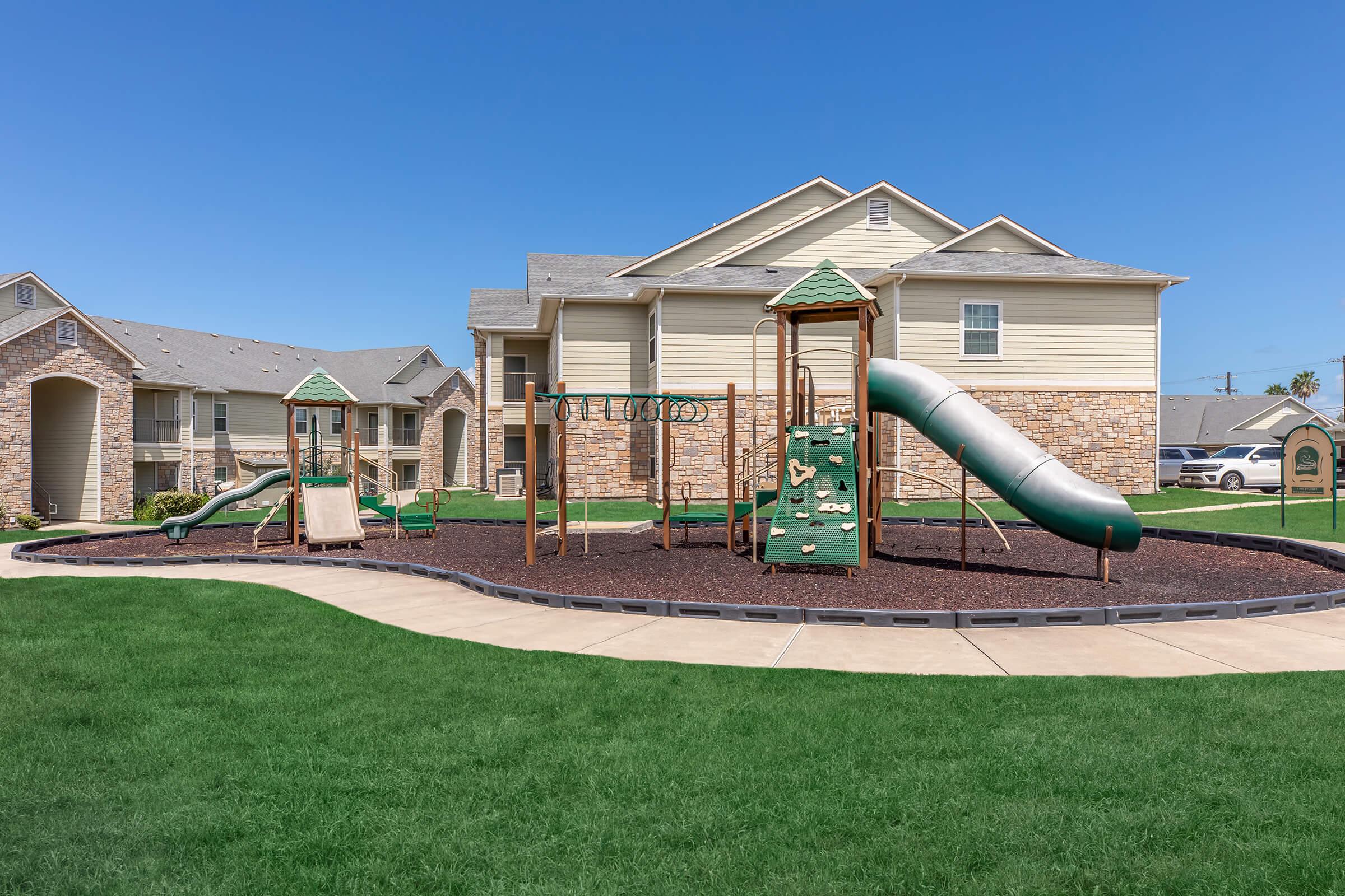 A sunny playground featuring a slide, climbing structure, and swings, surrounded by green grass and concrete pathways. In the background, there are residential buildings with beige siding and stone accents, under a clear blue sky.