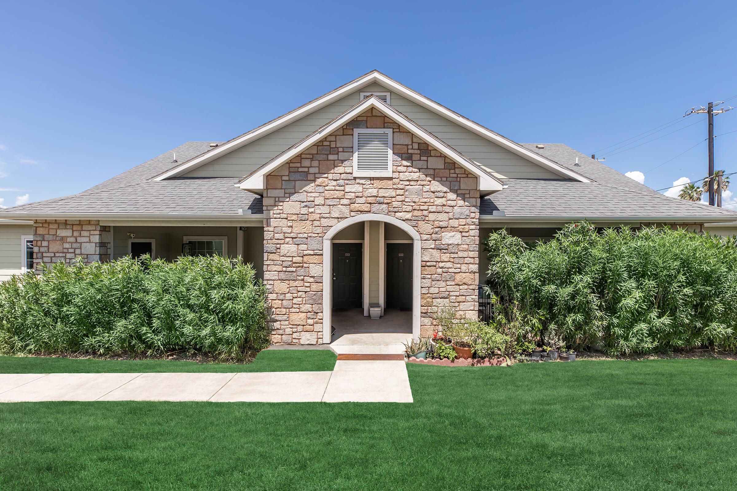 A charming single-story house with a stone facade, featuring an arched entryway. The front yard is well-maintained with lush green grass and bushy plants flanking the entrance. The sky is clear and blue, enhancing the home's inviting appearance.