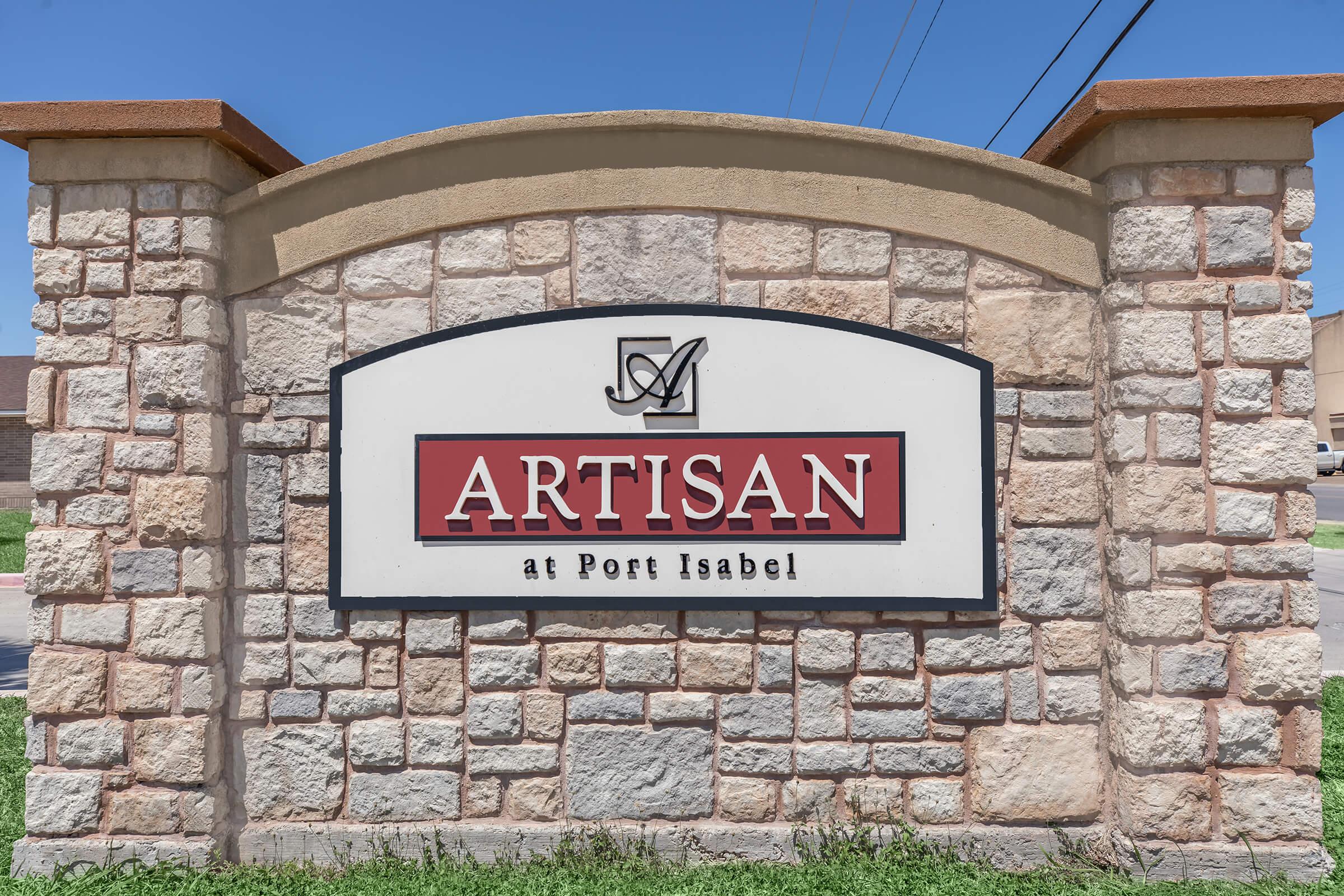 Sign reading "ARTISAN at Port Isabel" displayed on a stone structure, with a blue sky in the background. The sign features a decorative emblem above the text.