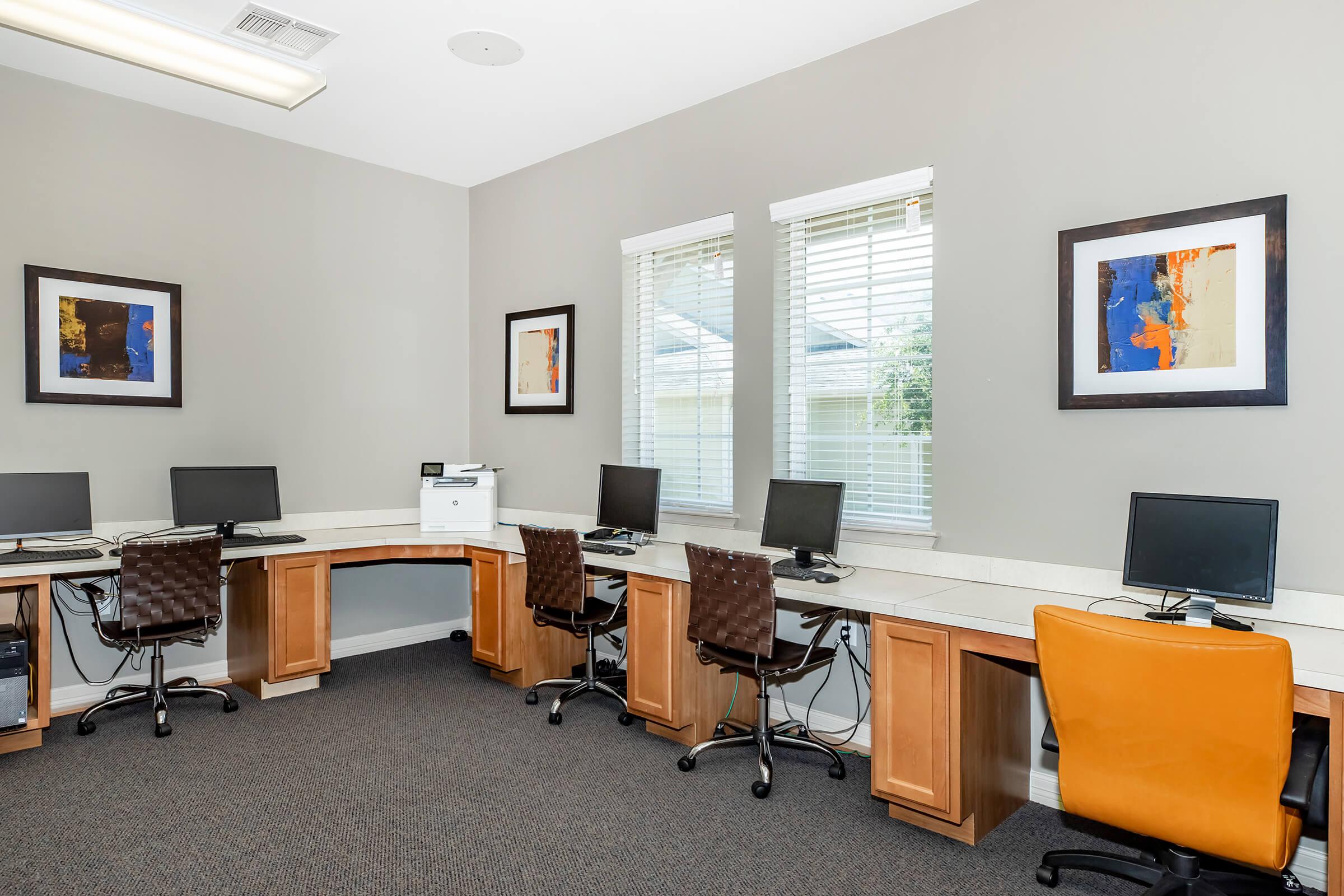 A modern workspace featuring several computer desks with chairs, two computers on each desk, and a printer. The walls are painted a neutral color and adorned with framed artwork. Natural light comes through two windows with white blinds, creating a bright and inviting atmosphere.