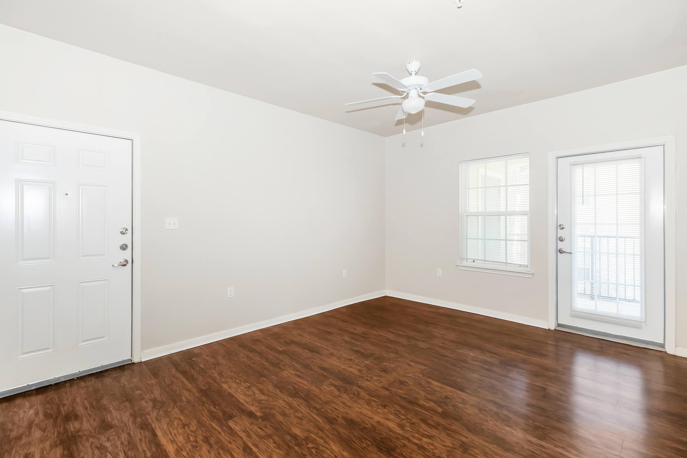 Bright, empty room featuring medium brown hardwood flooring, a white ceiling fan, and neutral walls. There is a front door on the left, a window on the right, and a door leading to a balcony. Natural light flows in through the windows, creating a spacious feel.