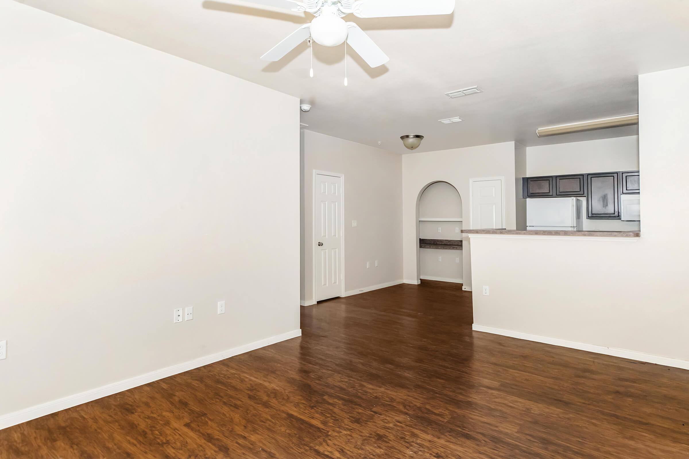 Spacious living area with light-colored walls and dark wood flooring. A ceiling fan hangs from the ceiling, and a kitchen is partially visible in the background through an arched opening. The space is empty, creating a bright and open feel ideal for home decoration.