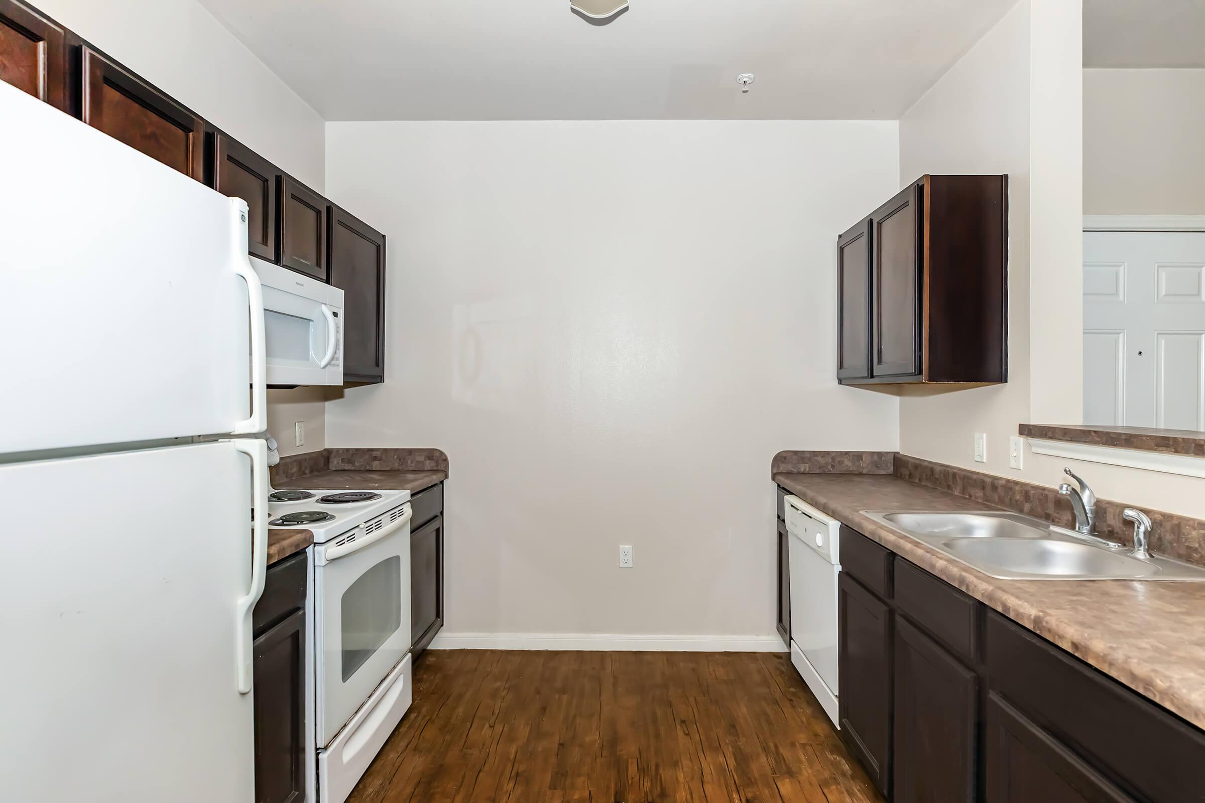 A kitchen featuring dark wooden cabinets, a white refrigerator, an oven, a microwave, and a dishwasher. The countertops are brown, and the floor is wooden. The wall is painted a light color, and the layout appears open and spacious.
