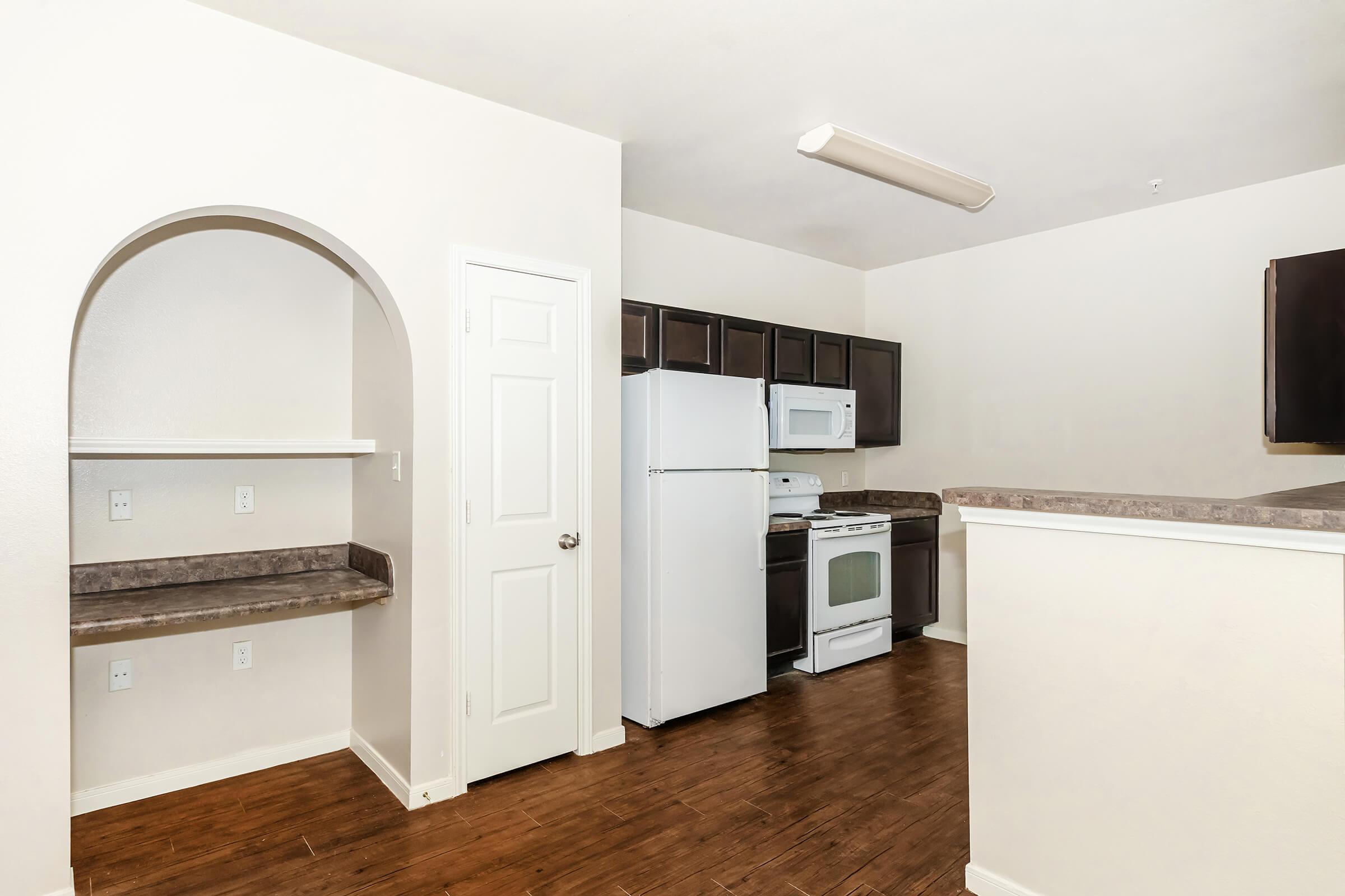 A modern kitchen featuring dark cabinetry, a white refrigerator, and a microwave. The space includes a stove and ample counter space, with a unique archway leading to an adjacent nook. The flooring is wooden, and the walls are painted in a light color, creating a bright and inviting atmosphere.
