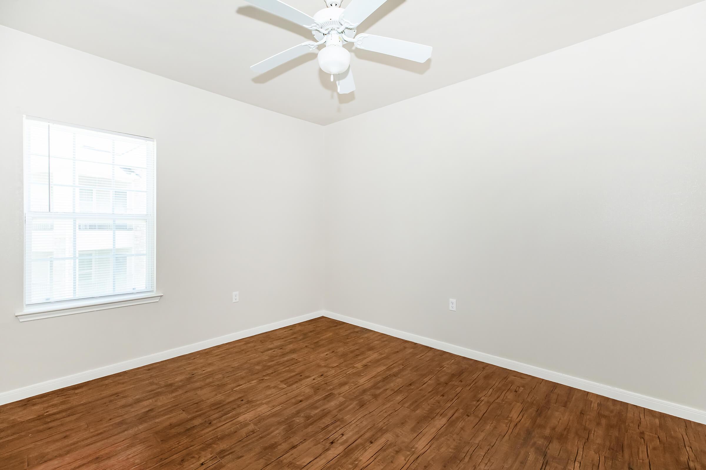 A bright, empty room with light-colored walls and a ceiling fan. The floor features warm-toned wooden planks, and there is a window that lets in natural light.