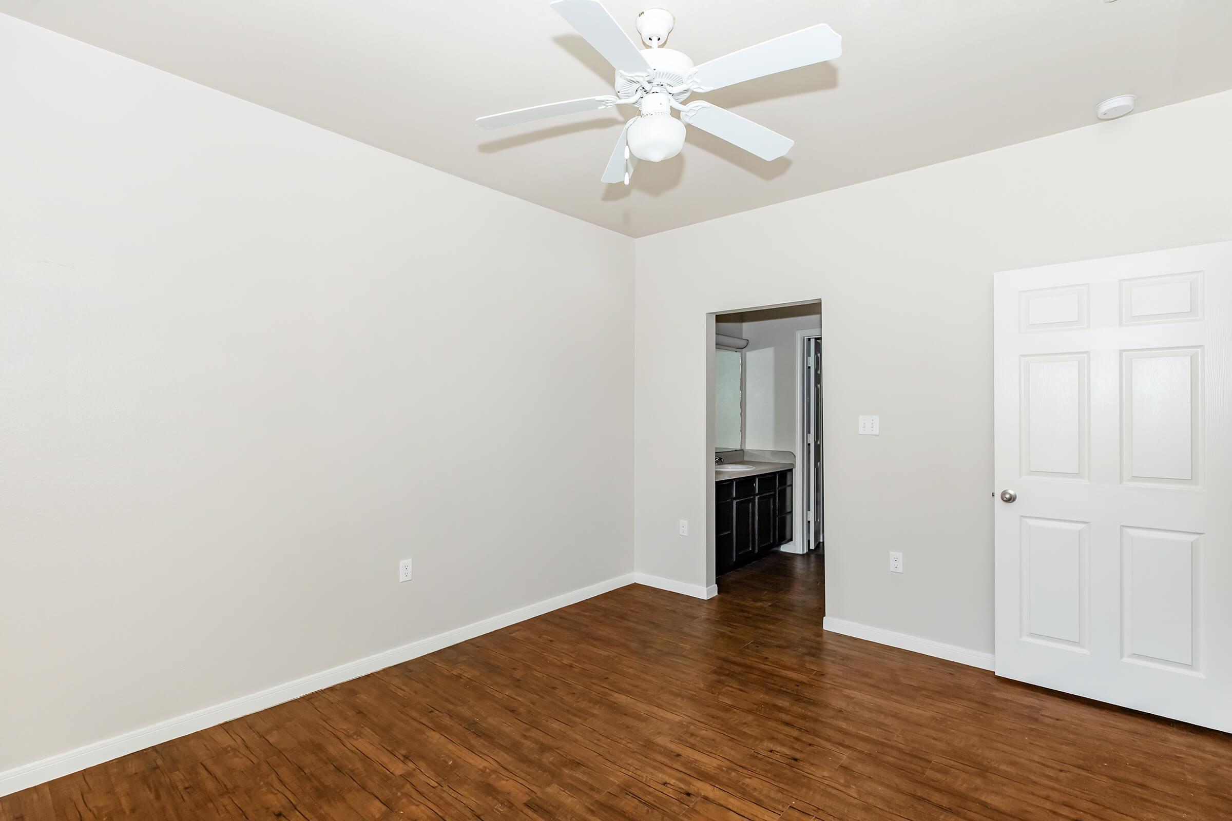 A well-lit, empty room with light beige walls and a wooden floor. A ceiling fan is visible. There is an open doorway leading to another room, and a closed white door is on the right side. The space appears clean and inviting, suitable for various uses.