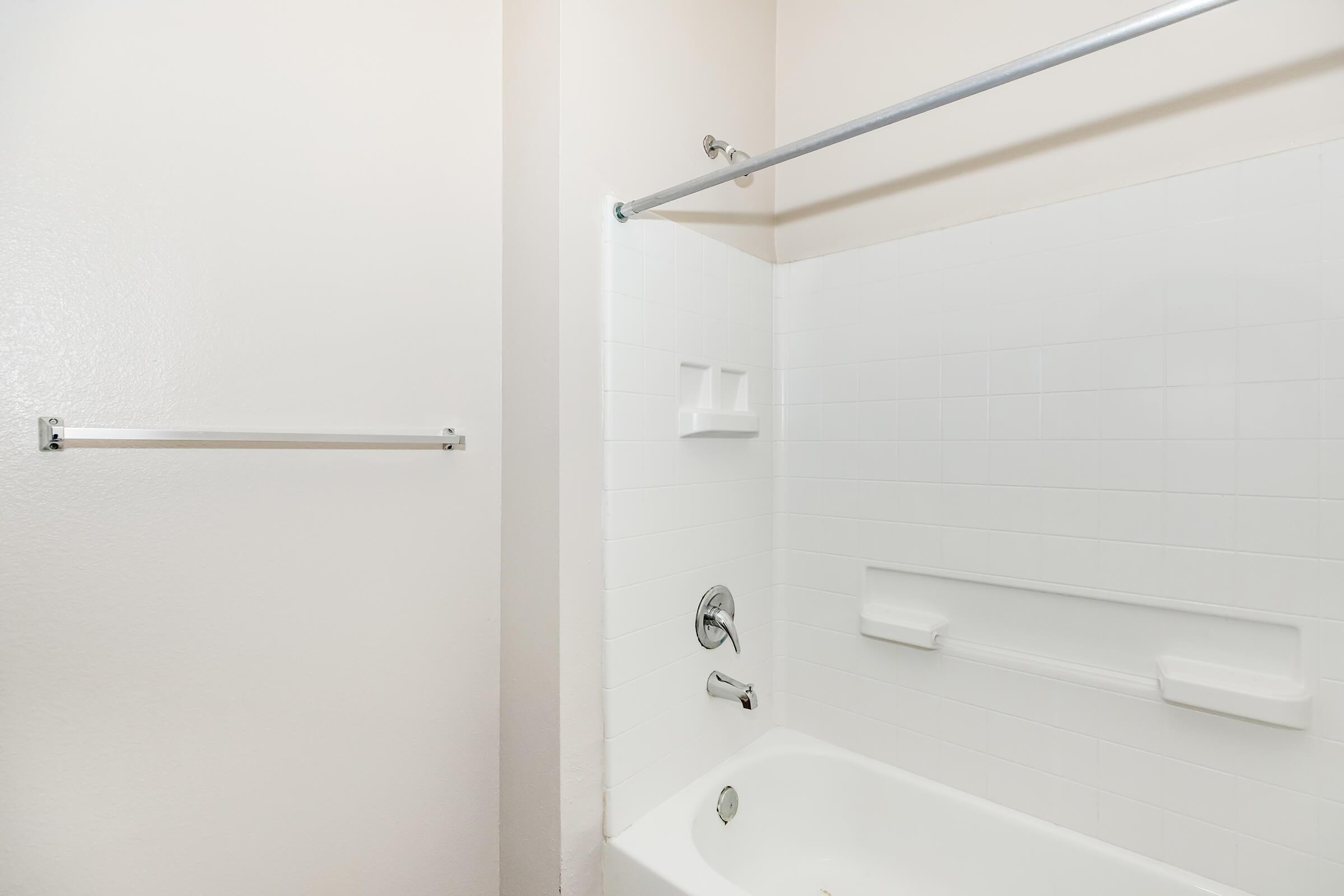 A clean bathroom featuring a white tiled bathtub and shower area. There is a chrome shower rod above the tub and a wall-mounted towel bar to the left. The walls are a light color, and the overall ambiance is simple and modern.