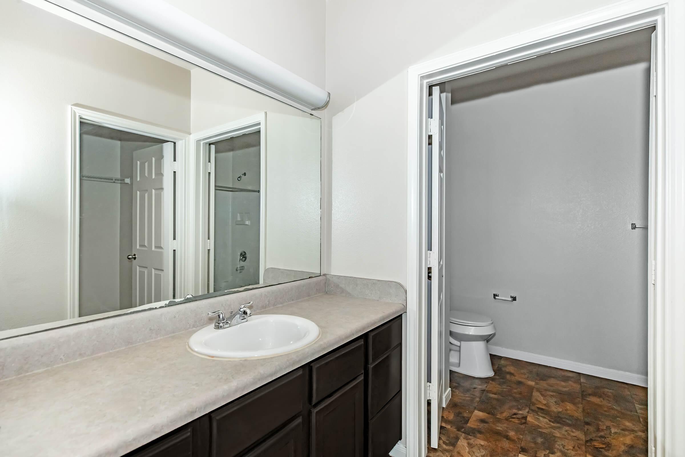 A well-lit bathroom featuring a double sink vanity with a light-colored countertop and dark wood cabinets. A large mirror hangs above the sink. To the right, an open door leads to a toilet area, with a simple tile floor and neutral-colored walls.
