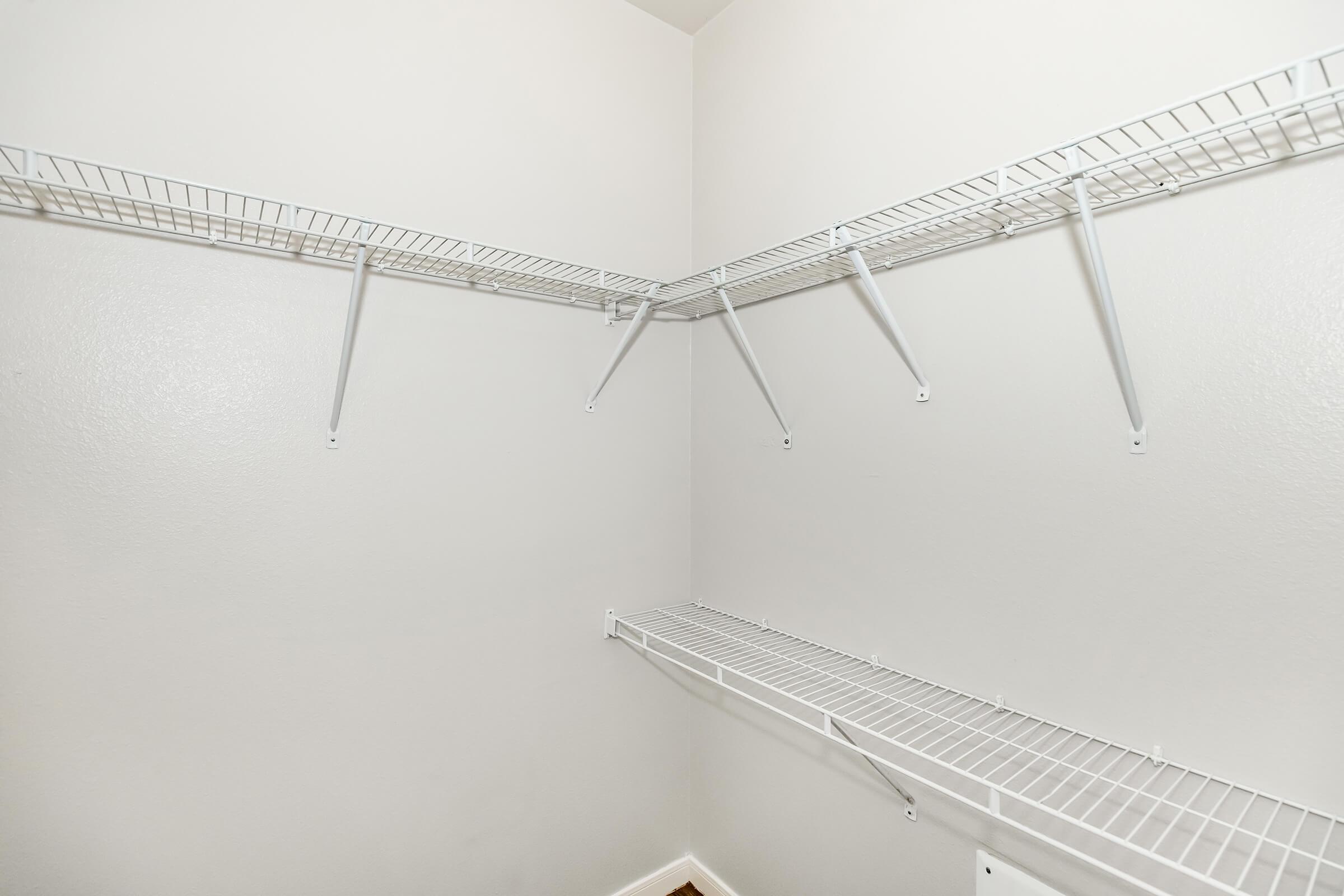 Empty corner closet with white wire shelving units attached to light-colored walls, providing a simple and functional storage solution. The floor is bare, highlighting the spaciousness of the closet. No items are stored on the shelves.