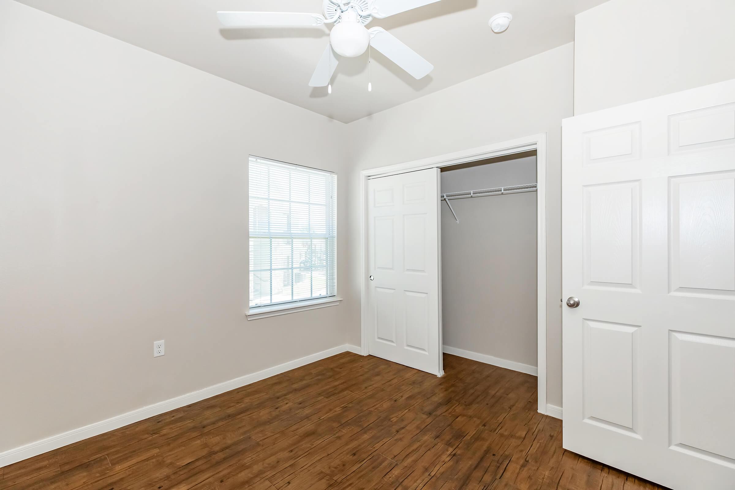 A bright, empty bedroom featuring light-colored walls and a wooden floor. There is a ceiling fan, a window with white blinds, and a closet with double doors on one side. The room is spacious and well-lit, providing a clean and minimalist look.