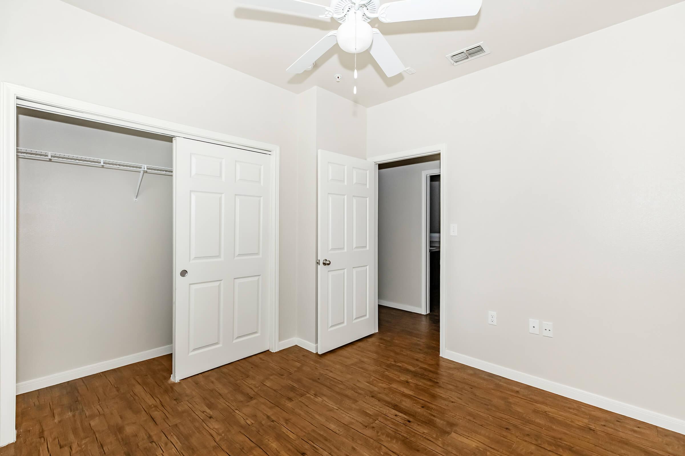 A spacious, well-lit room with light-colored walls and hardwood flooring. There are two closed doors on the left side and another door leading to a darker area in the background. A ceiling fan is visible, and a closet with a wire shelf is present on the left. The overall atmosphere is clean and uncluttered.