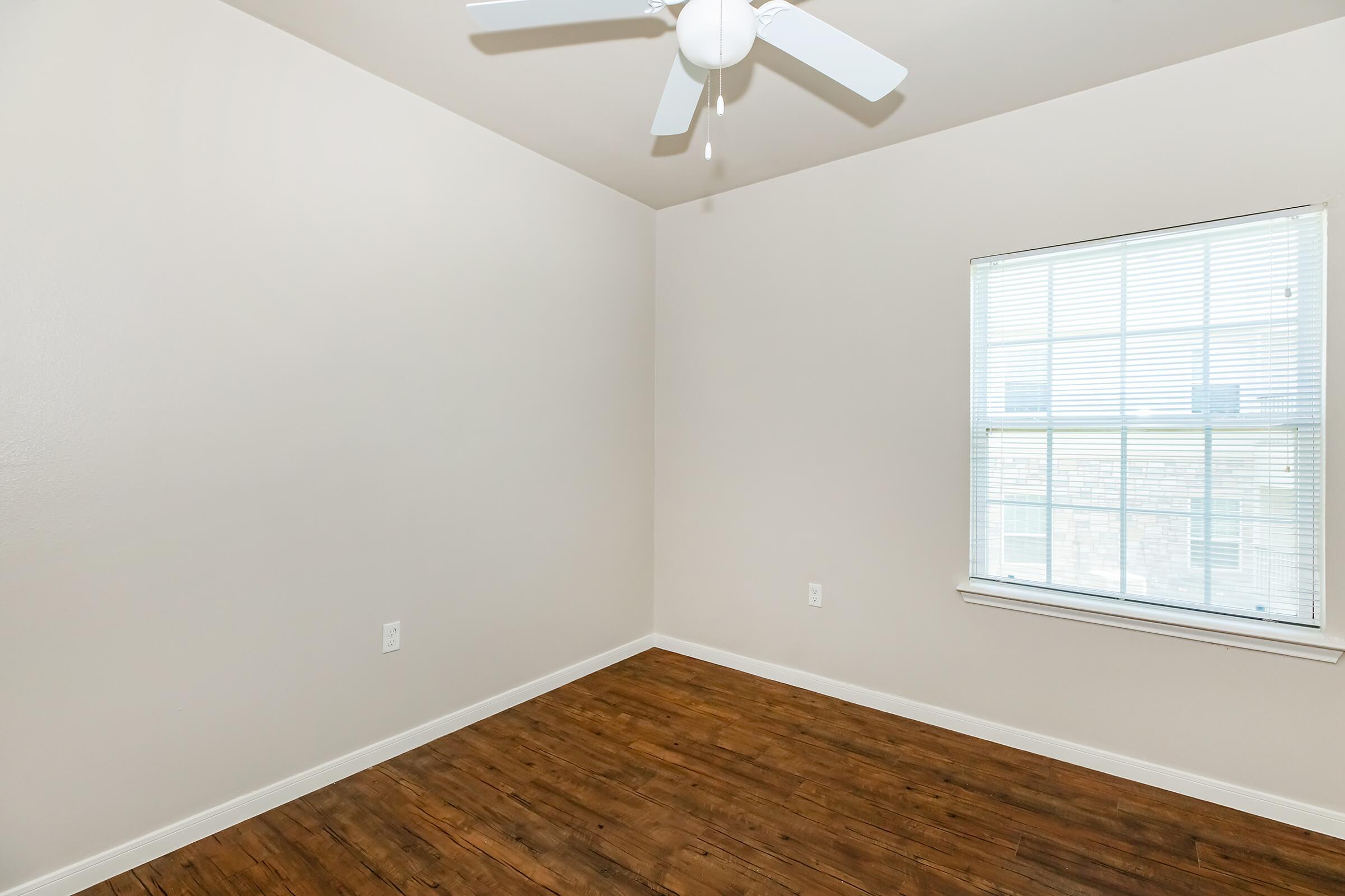 A minimalist room featuring light beige walls and wooden flooring. There is a ceiling fan with three blades and a large window with white blinds, allowing natural light to enter. The room is empty, emphasizing its spaciousness and neutral decor.