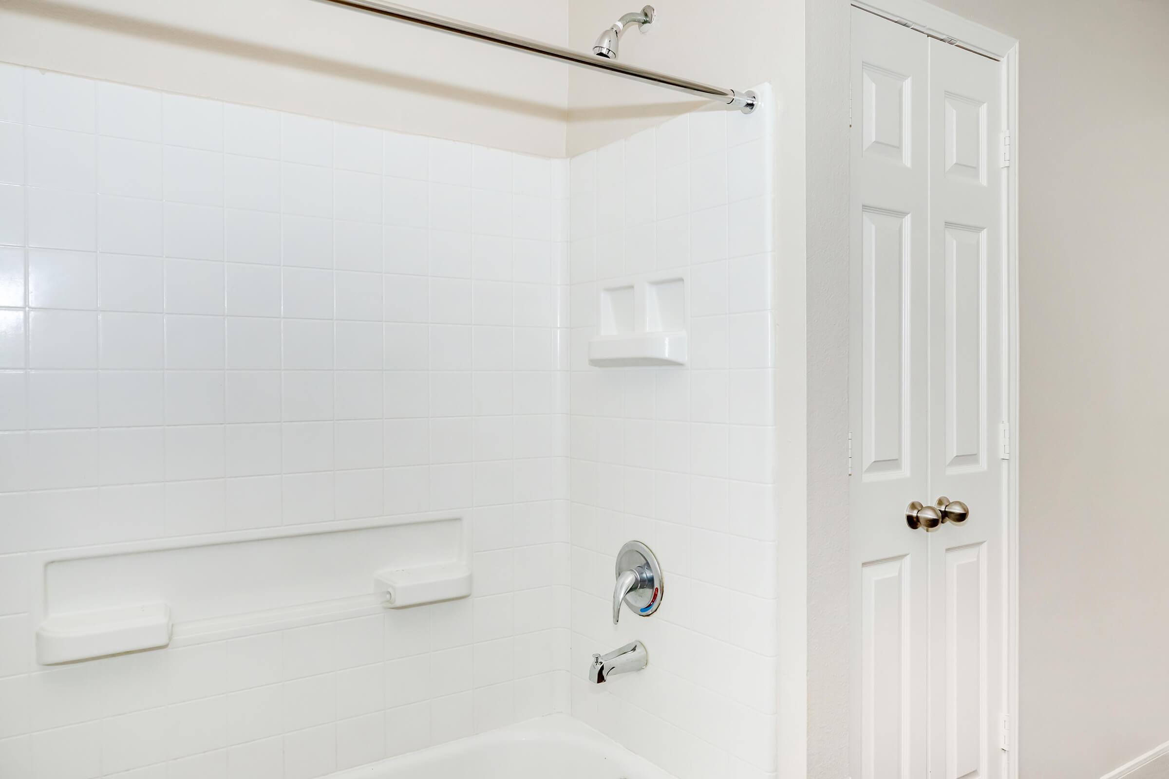 A clean, well-lit bathroom featuring a white tiled bathtub with a shower curtain rod. Two built-in shelves are visible on the wall above the tub, and a chrome faucet is installed. A white door is partially visible on the right side, providing access to another space.