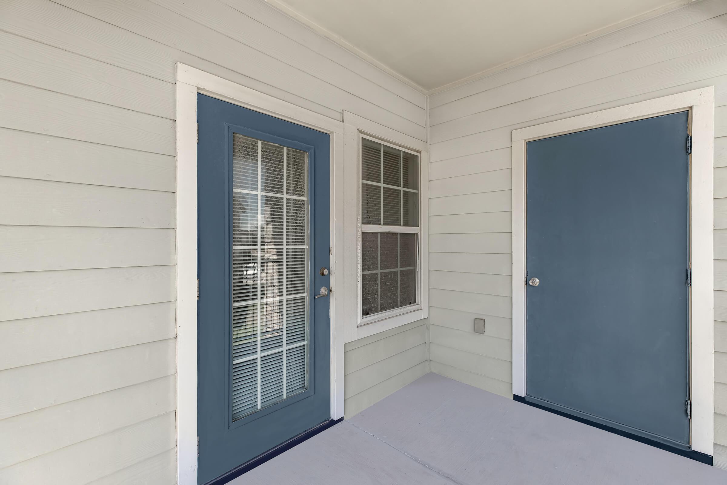 A porch area featuring two doors. The left door has a window with crisscrossed grids, while the right door is solid. Both doors are painted blue, and the walls are light gray siding. The floor is a neutral shade, adding to the welcoming entrance. Natural light is visible through the window.