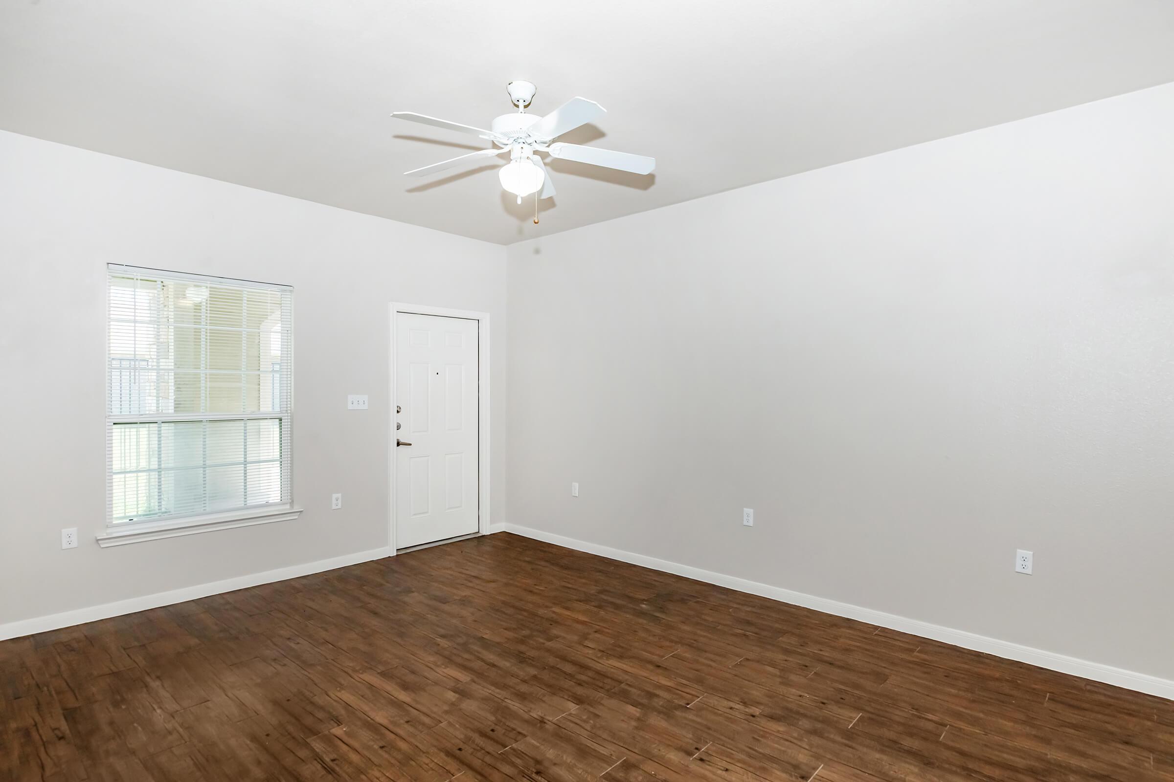 A spacious, empty room featuring a ceiling fan, light fixtures, and a large window. The walls are painted light colors, and the floor has a wood-like finish. A front door is visible along one wall, creating a bright, airy atmosphere in the space.