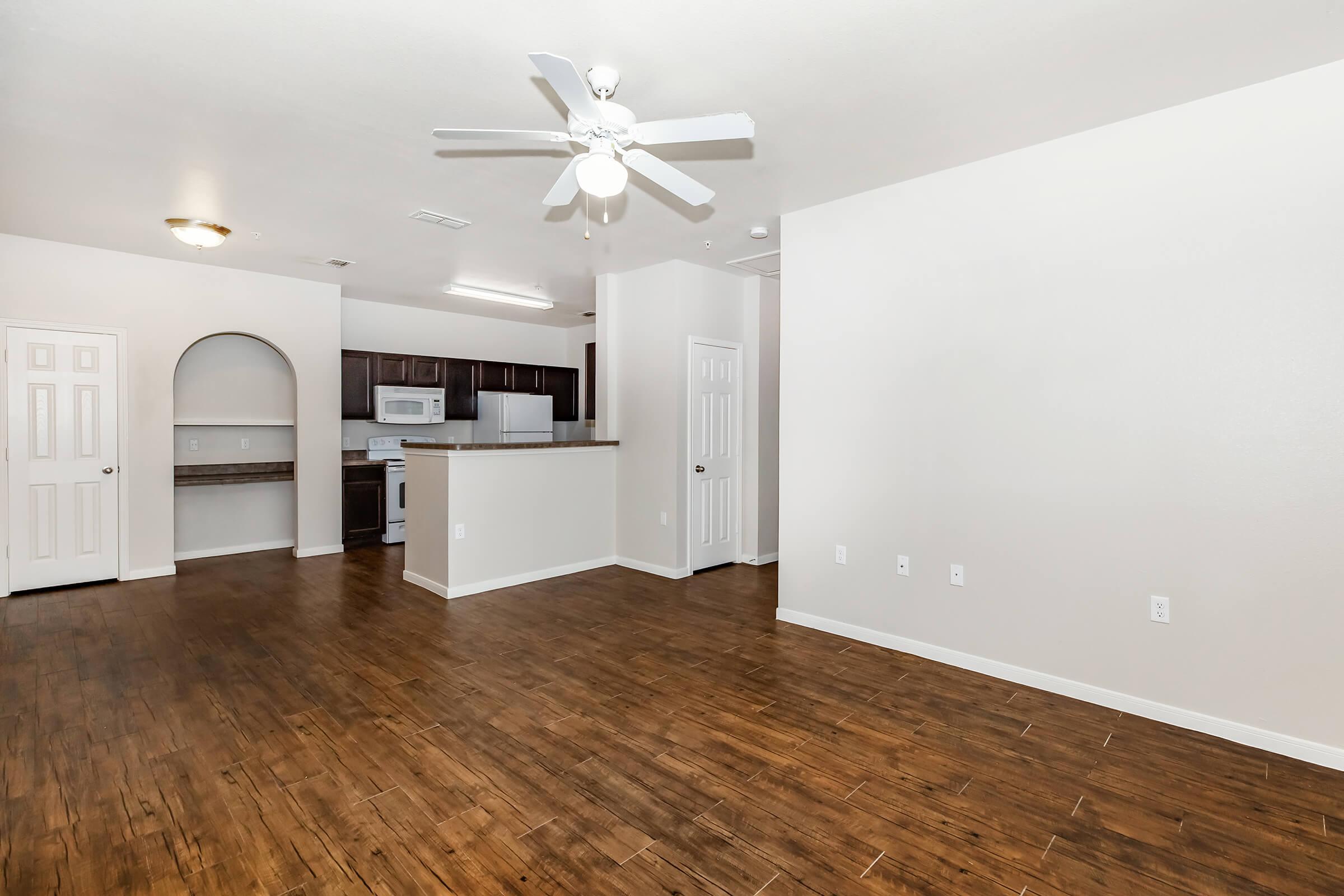Spacious interior of an apartment featuring a living area with hardwood floors, a ceiling fan, and neutral-colored walls. The open kitchen is visible in the background, equipped with dark cabinetry and modern appliances. There are doors leading to other rooms and a counter area for breakfast.