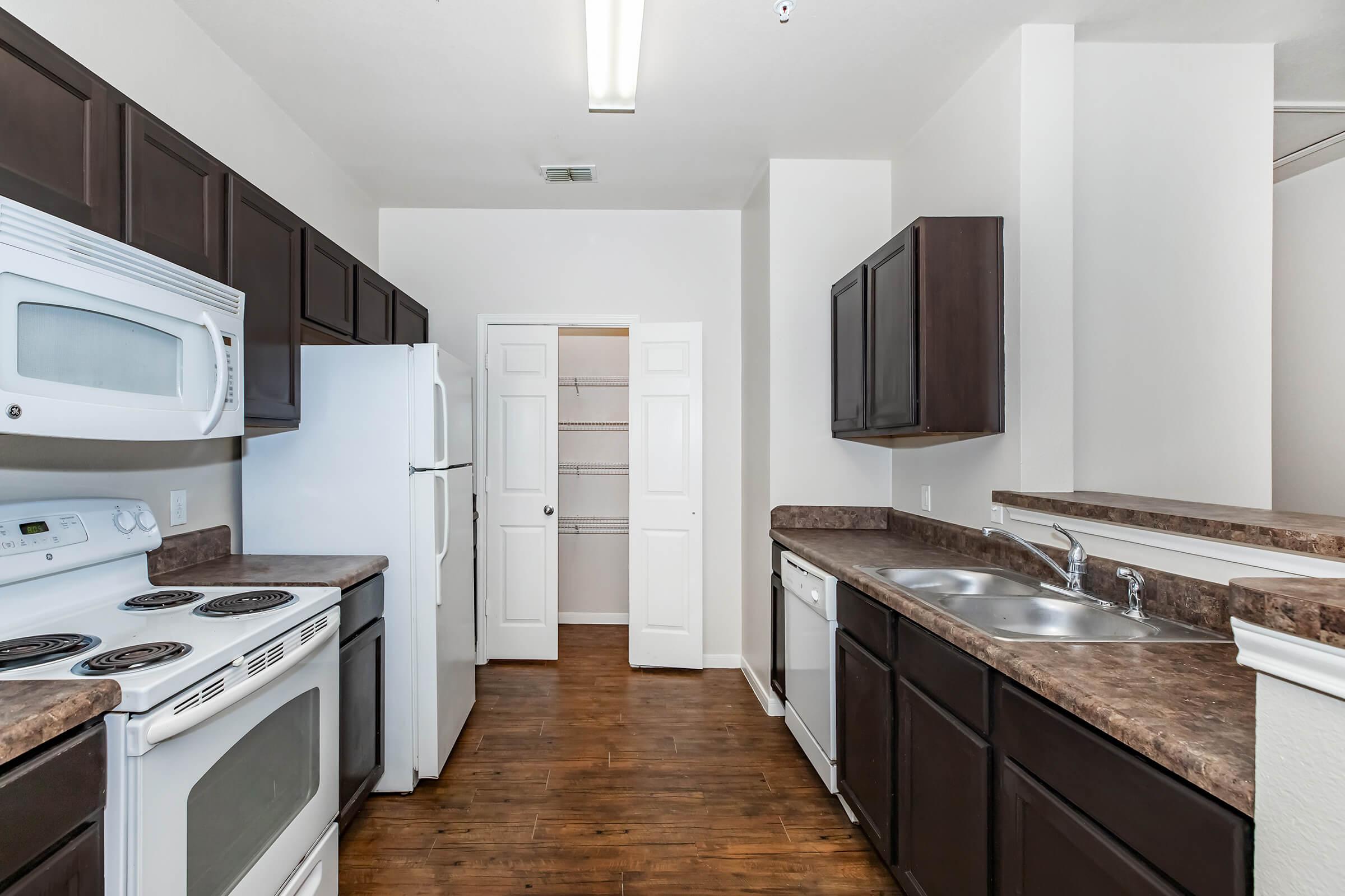 A modern kitchen featuring dark wood cabinetry, white appliances including a refrigerator, stove, and dishwasher. The countertop is dark with a subtle texture. A pantry closet is visible through an open door. The flooring is a warm wood-like material, and the space is well-lit with overhead lighting.