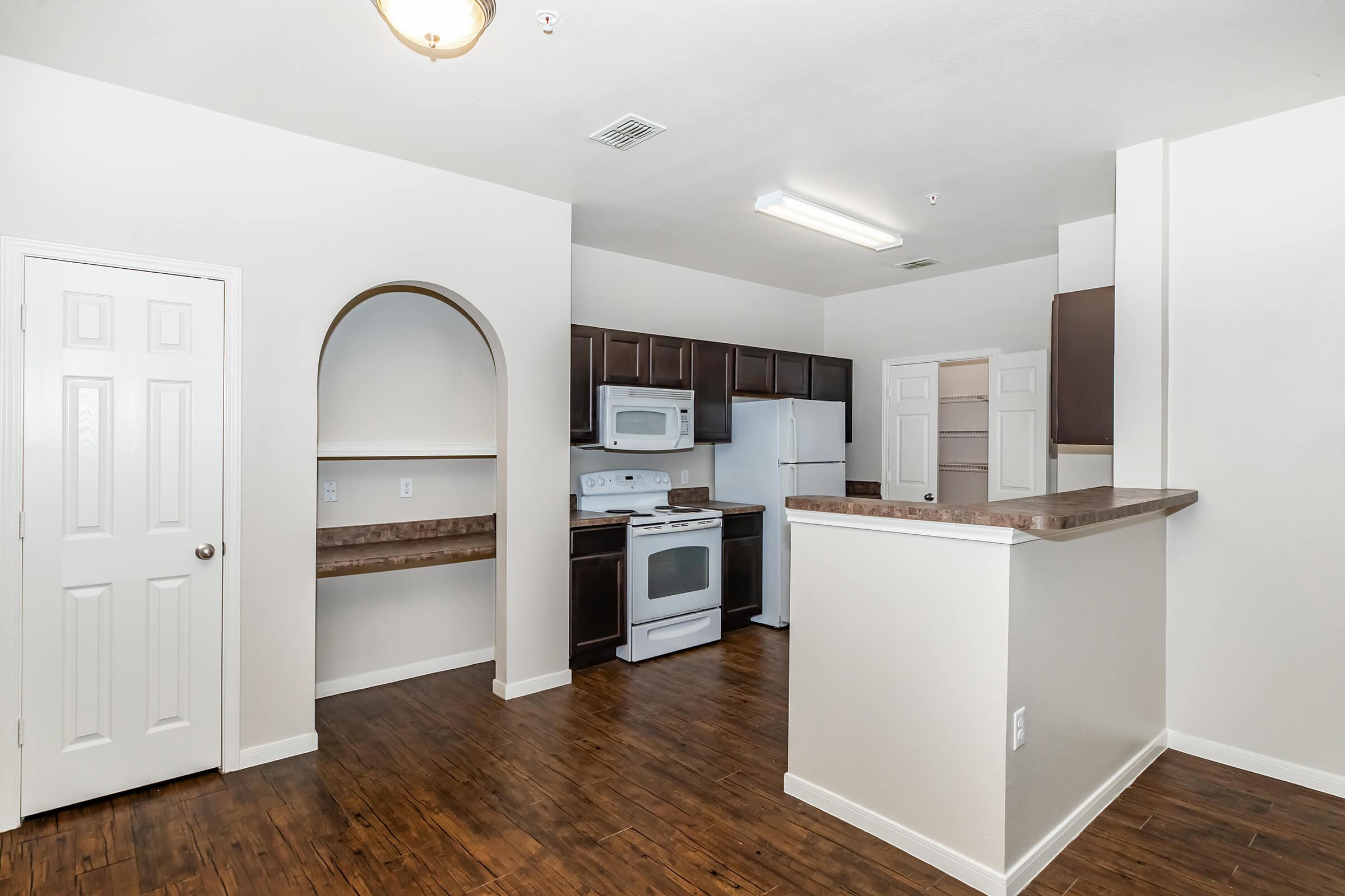 A modern kitchen and living space featuring dark cabinetry, a white stove and refrigerator, and wooden flooring. The kitchen includes an open layout with a bar area and a small pantry, alongside a door leading to a room. Bright overhead lighting enhances the airy atmosphere.