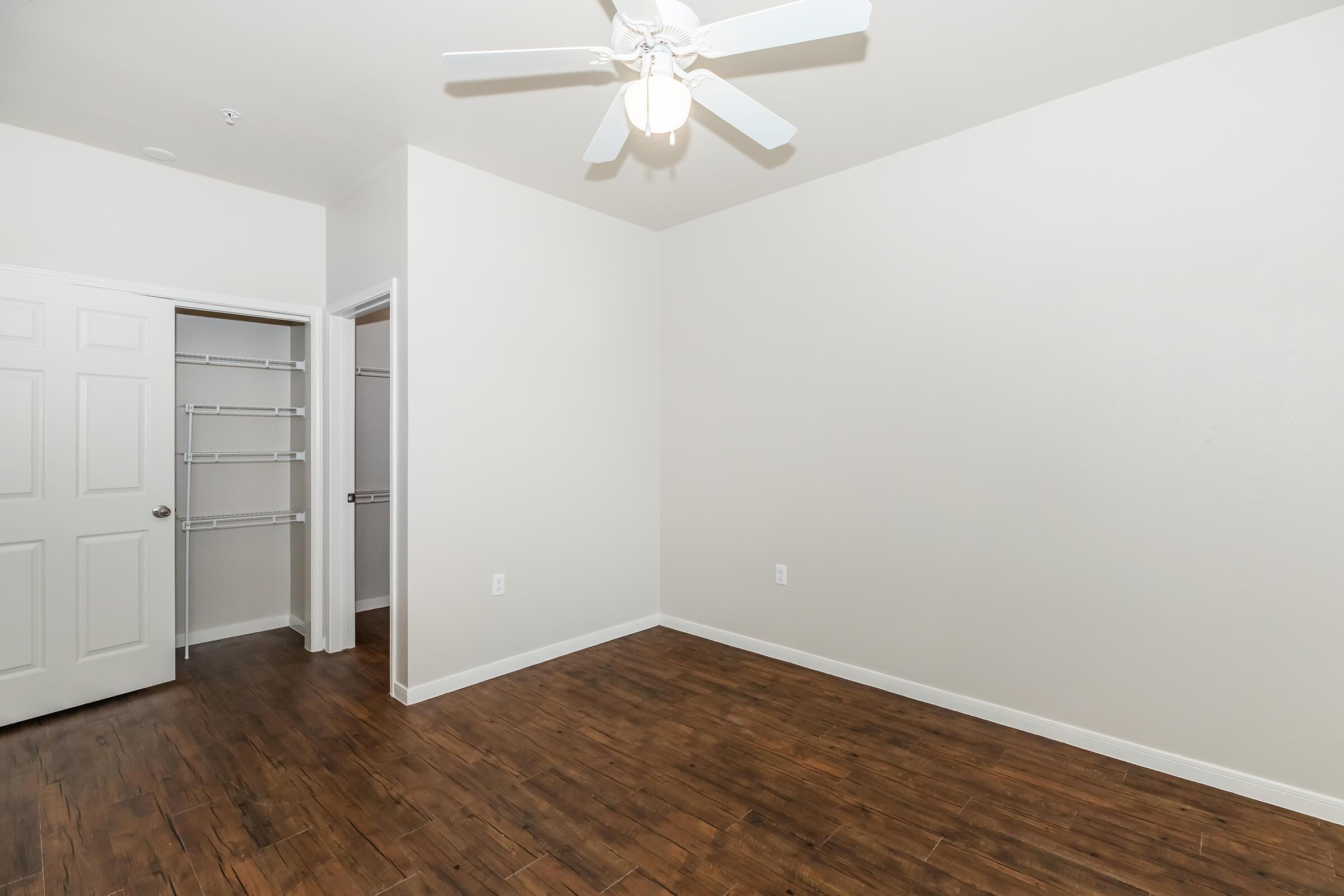 A spacious room with a light-colored wall and wooden flooring. It features a ceiling fan and an open closet with shelves. One wall has a door leading to another room, showcasing a minimalist design and ample natural light.