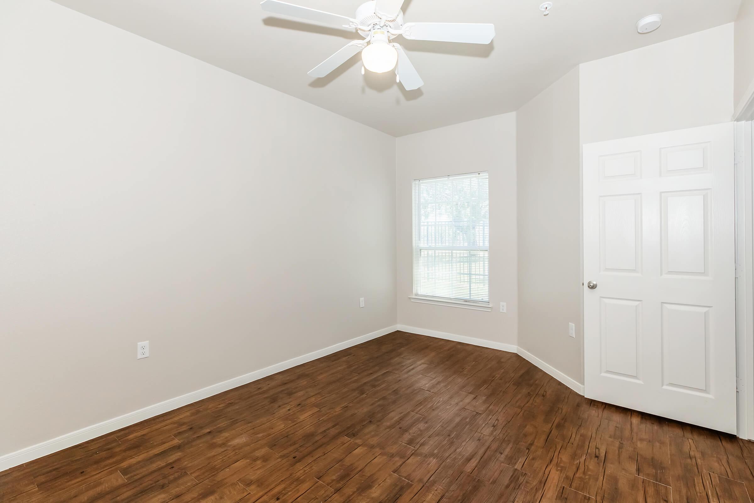 A spacious, empty room with light-colored walls and a wooden floor. It features a ceiling fan, a window allowing natural light, and a closed white door. The room has a minimalist design, making it suitable for various uses such as an office or bedroom.
