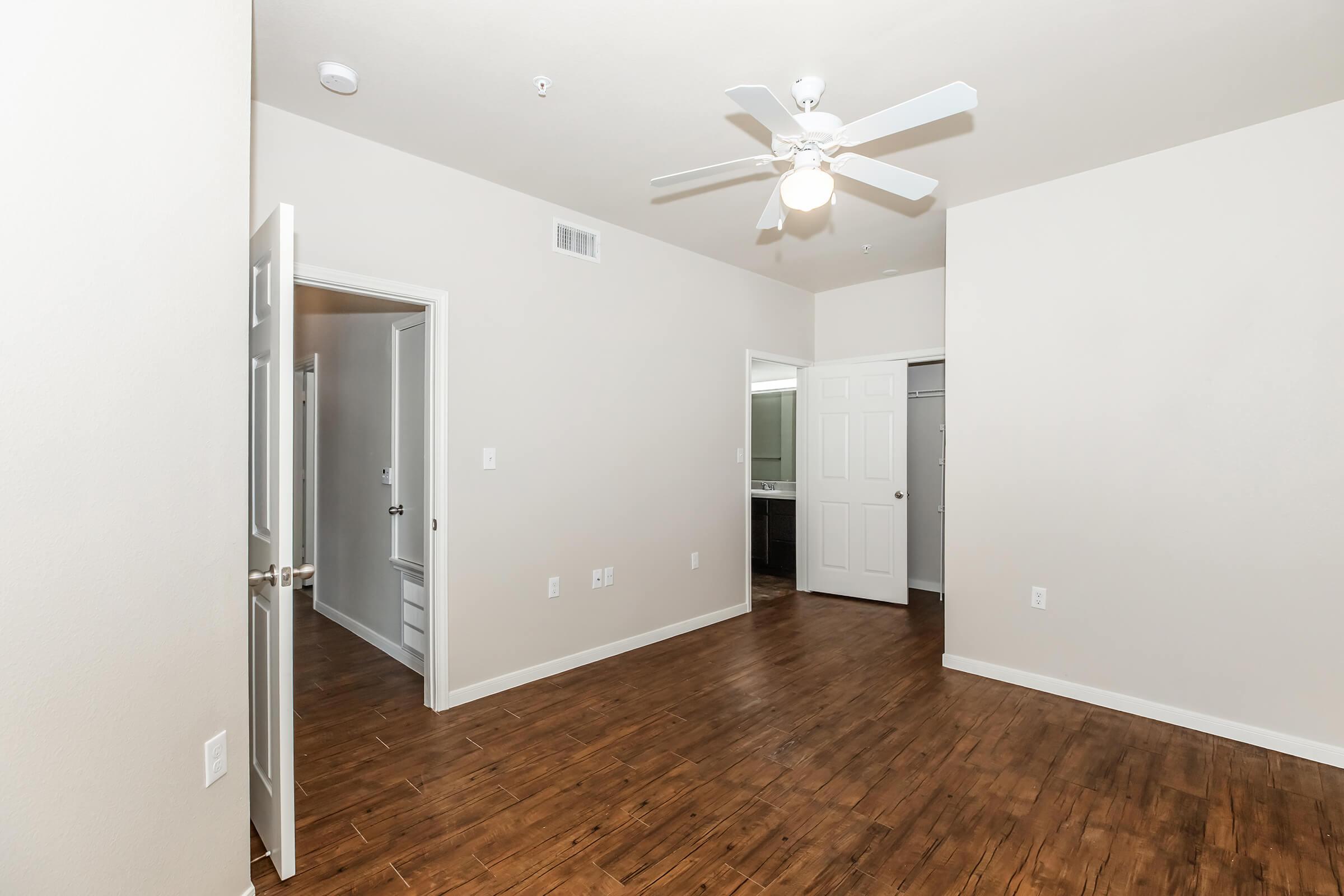 Spacious interior of a room featuring light-colored walls, a wooden floor, and a ceiling fan. Doorways lead to adjacent rooms, with one door open to a closet. The overall ambiance is bright and inviting, ideal for various interior design options.