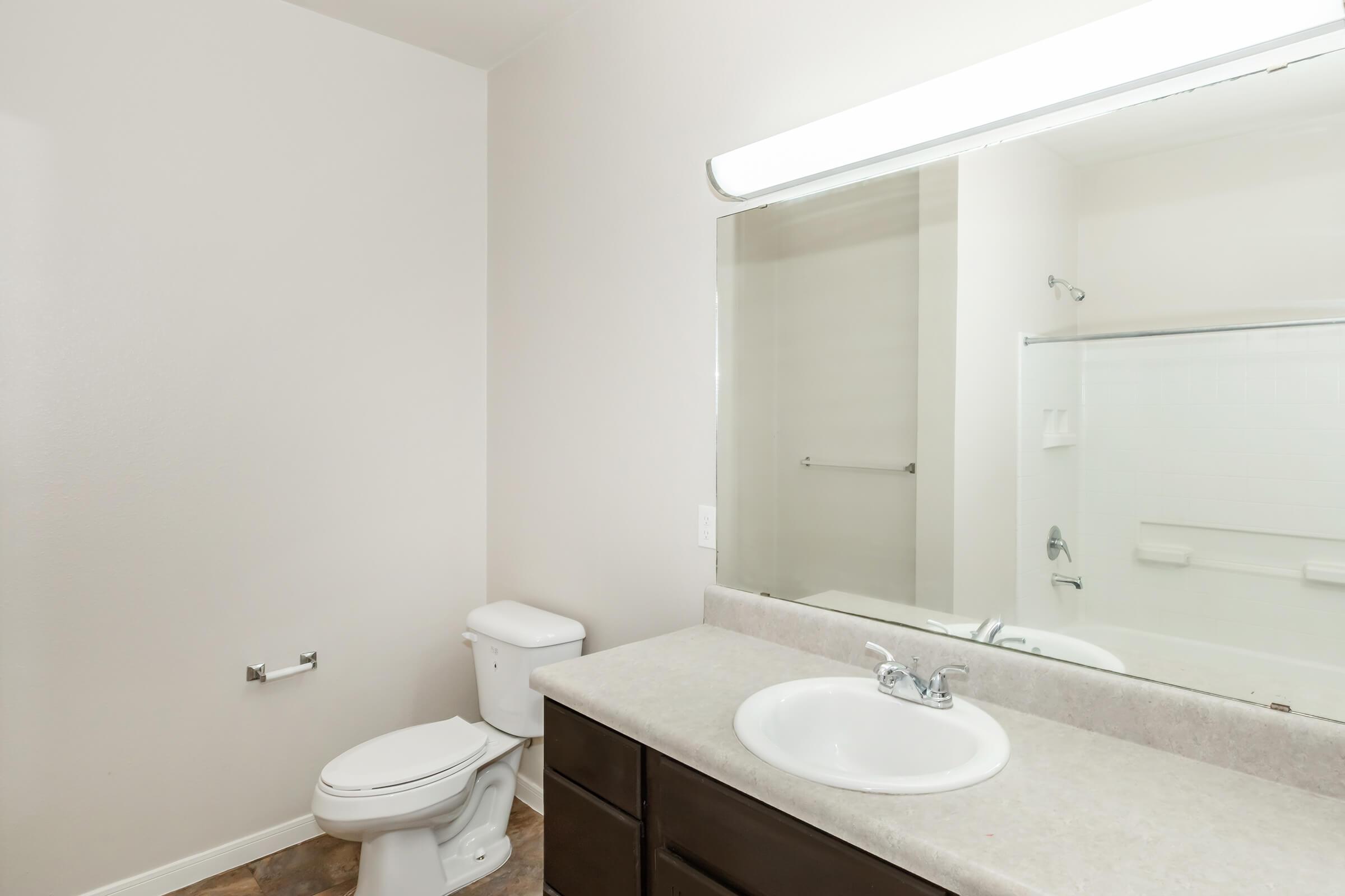 A clean and modern bathroom featuring a white toilet, a sink with a countertop, a large mirror, and a bathtub with a shower curtain. The walls are painted in a light color, and the floor has a neutral tone. Bright lighting is provided by a LED light fixture above the mirror.