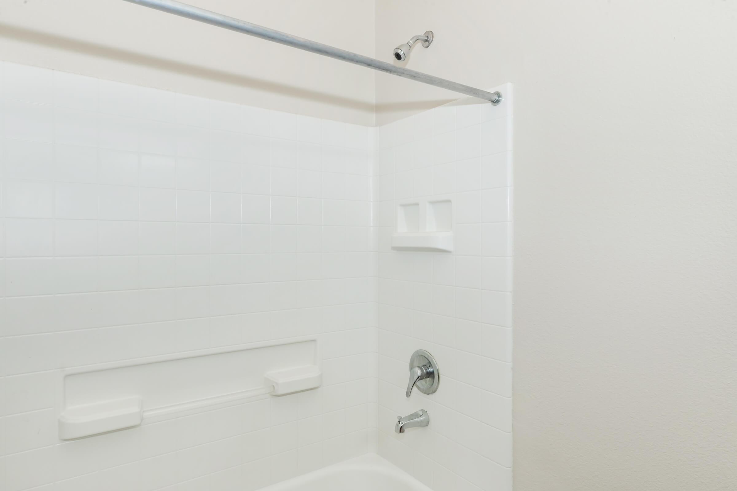 A clean, modern shower stall featuring white tiles, a showerhead, and a chrome shower rod. The walls include built-in shelves for toiletries, and the space is well-lit with neutral-colored walls. The overall design is simple and functional.