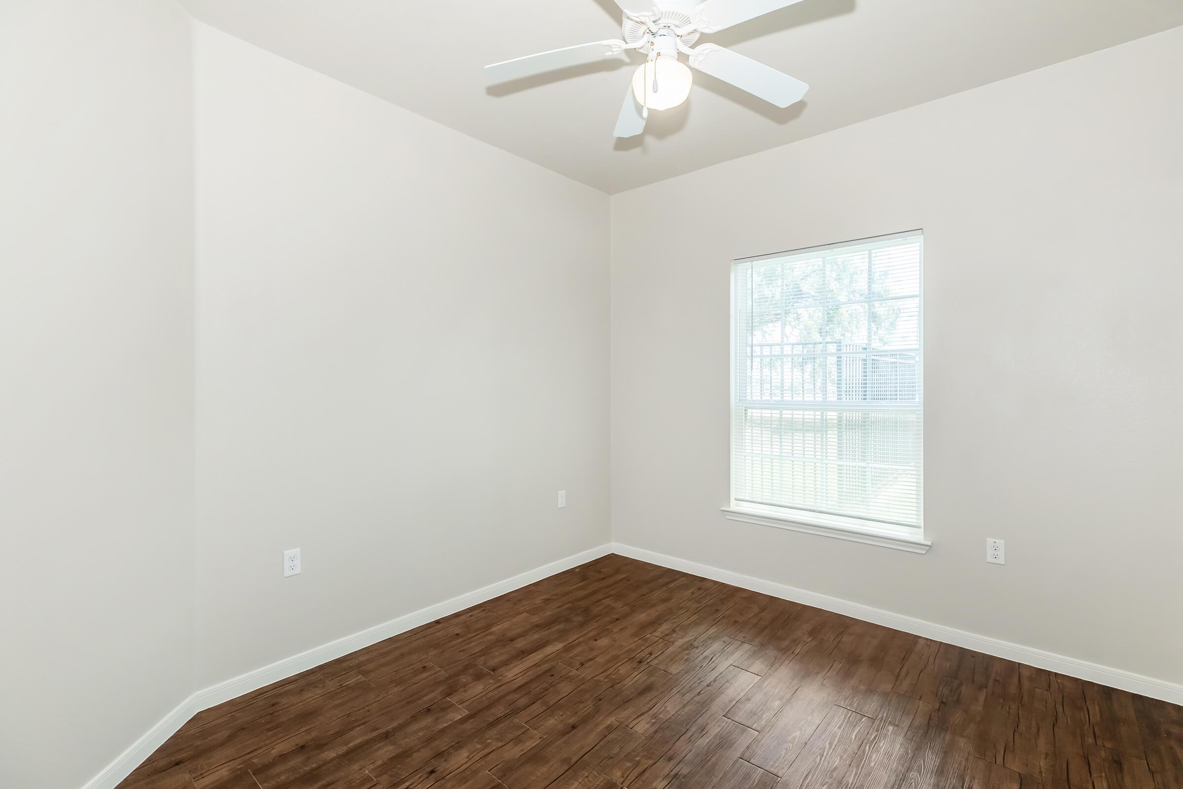 A bright, empty room featuring light-colored walls, a ceiling fan, and a large window allowing natural light to enter. The floor has a wood-like finish, and the overall space is uncluttered, creating an inviting atmosphere.