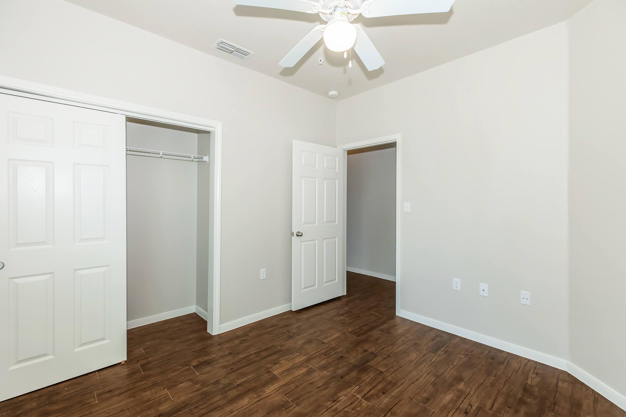A well-lit, empty room featuring light-colored walls and a wood-style floor. Two doors lead to a closet and another space, while a ceiling fan is mounted on the ceiling. The room has a simple and clean design, suitable for various uses.