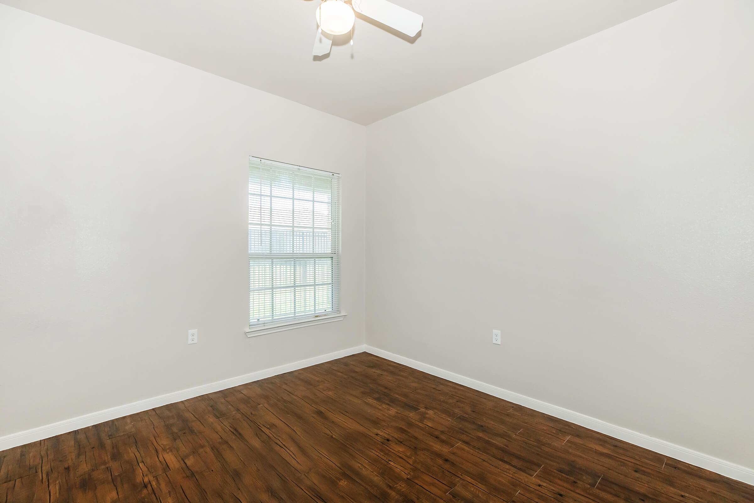 Empty room with light-colored walls and a wooden floor. There is a window with blinds allowing natural light to enter. A ceiling fan is visible, and the room appears to be well-lit and spacious, suitable for various uses.