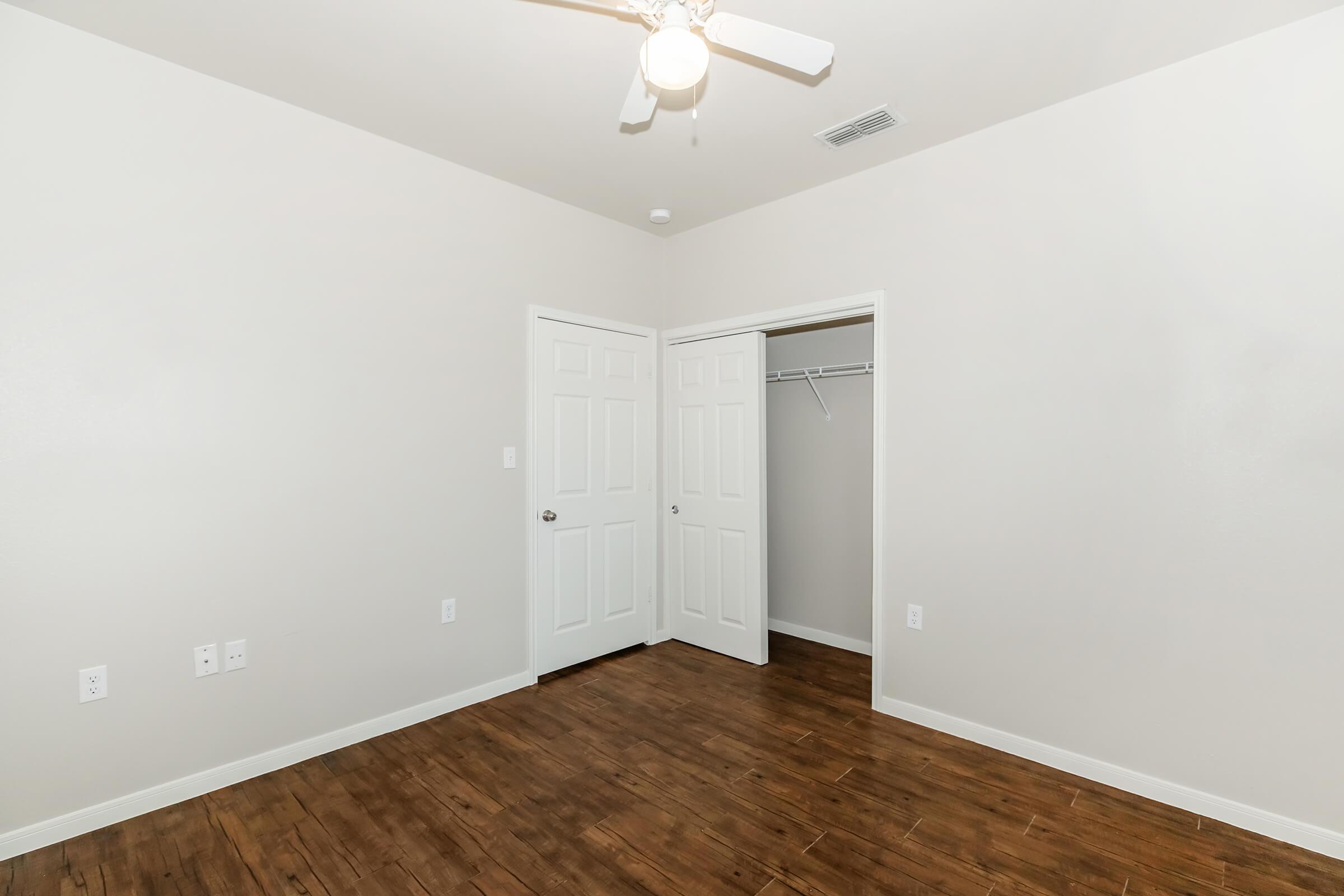Empty room with light-colored walls and a wooden floor. The room features a ceiling fan, two white doors leading to closets, and a small closet with no door. Natural light illuminates the space, creating a bright and open atmosphere.