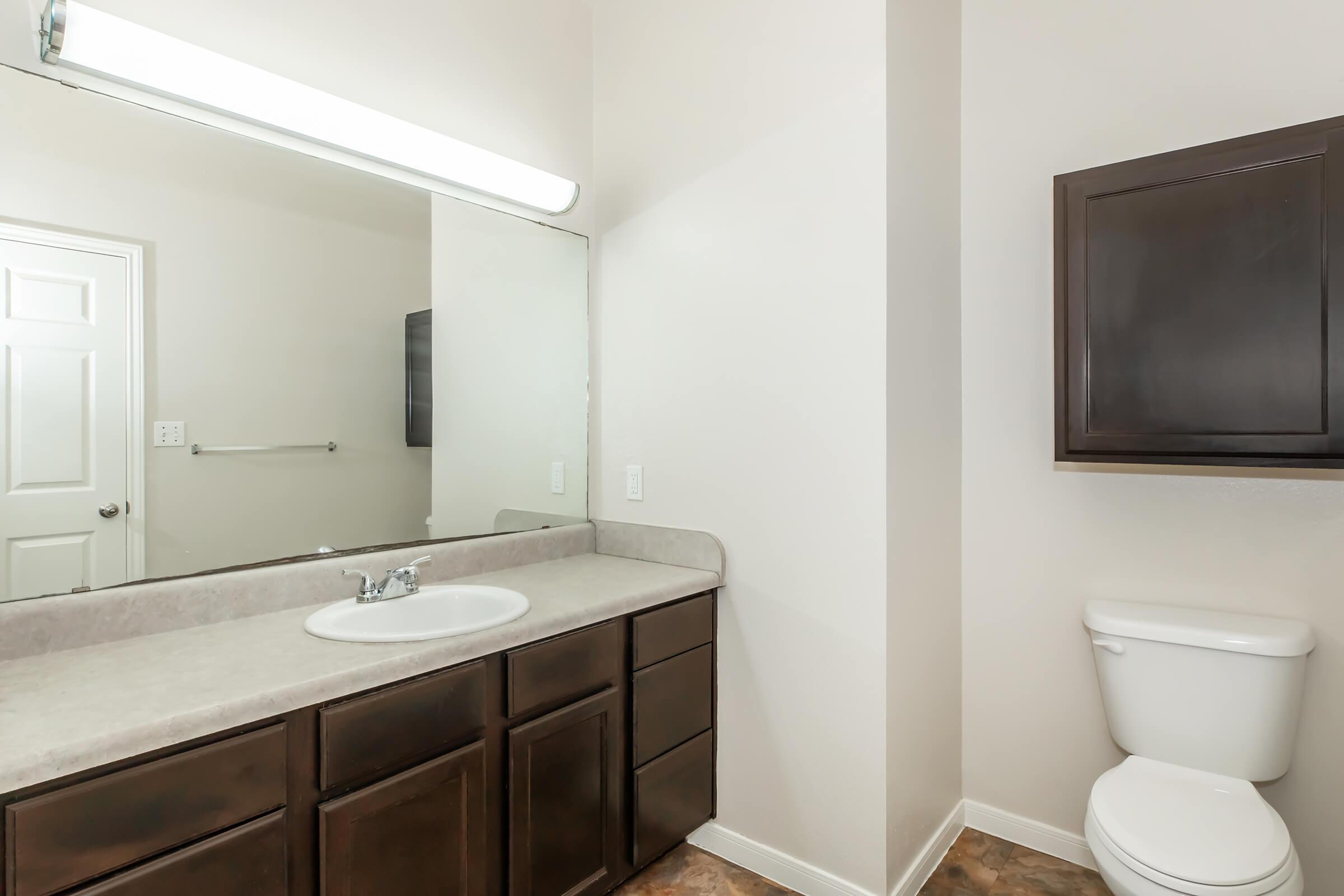 A modern bathroom featuring a large mirror above a double sink with dark wooden cabinets, a toilet, and a light-colored wall. The flooring has a pattern, and there is a wall-mounted cabinet next to the sink area. Bright lighting enhances the spacious feel of the room.