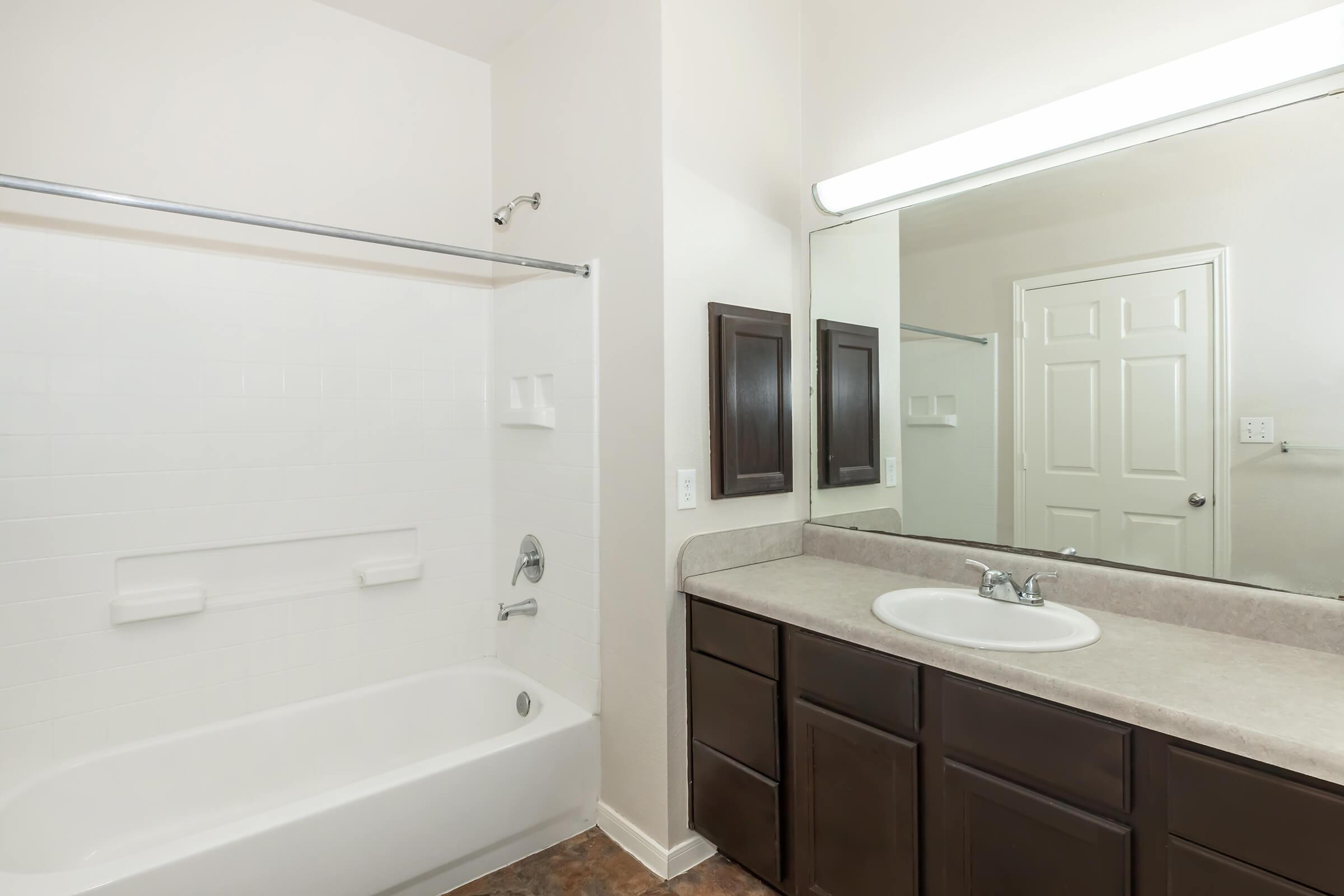 A clean and modern bathroom featuring a white bathtub with a shower curtain, a double sink vanity with dark wooden cabinets, and a large mirror. The walls are light-colored, and there is a door leading to another room. Soft lighting highlights the space.