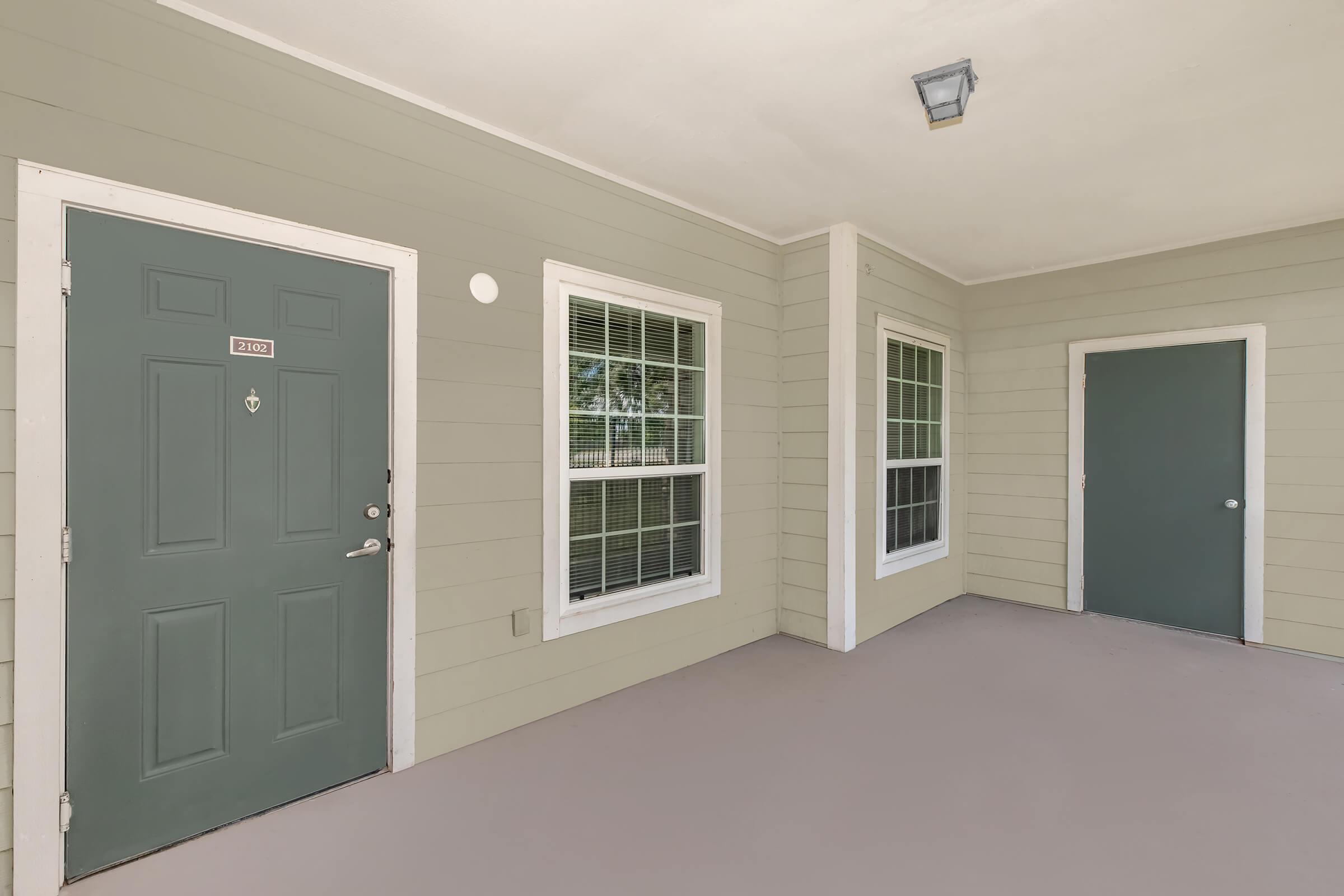 Exterior view of an apartment entrance featuring a green front door, two windows with white frames, and a second door on the right. The walls are painted light green, and the floor is a light-colored surface. A ceiling light fixture is visible overhead.