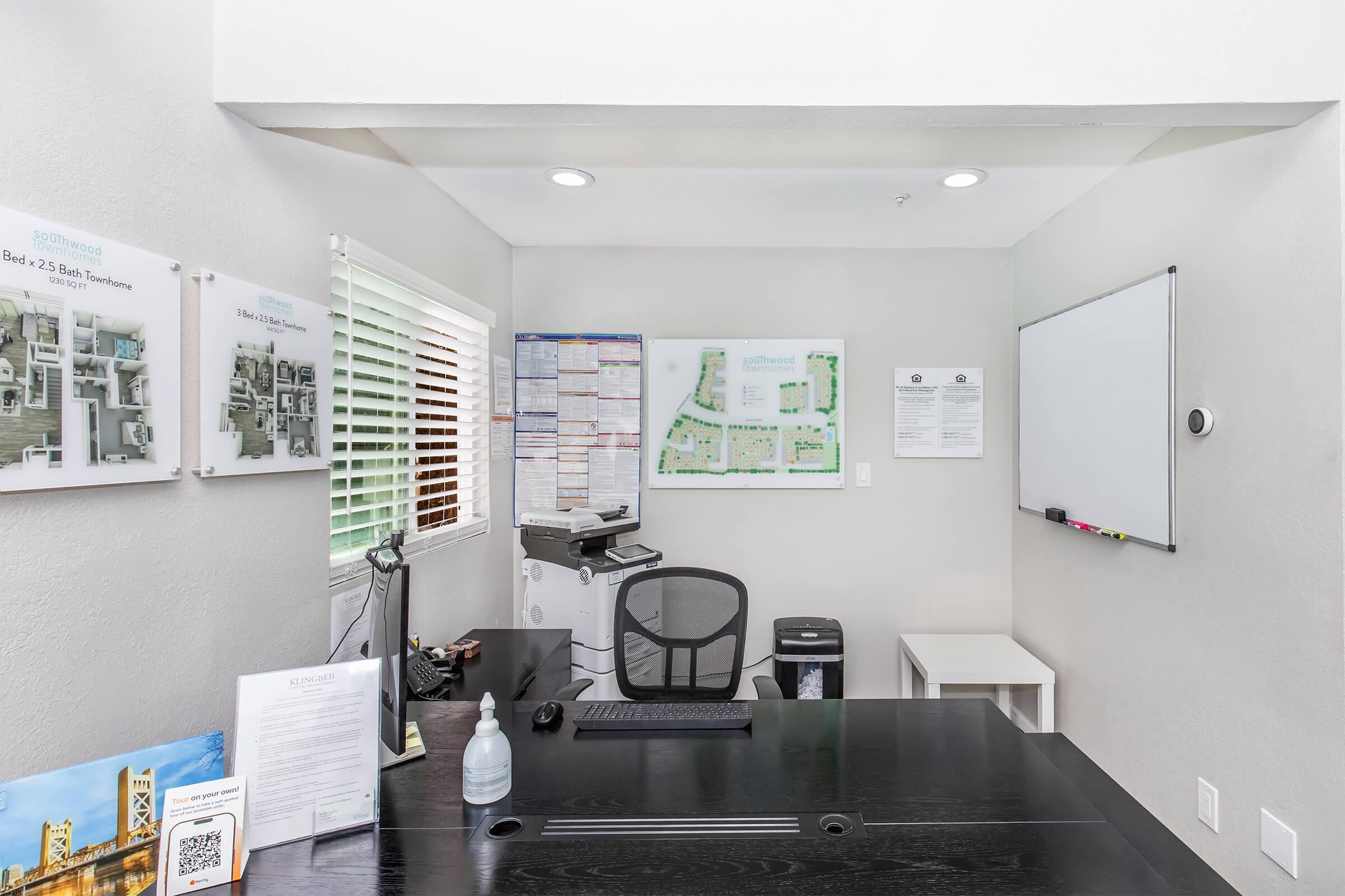 An office space featuring a black desk with a chair, a whiteboard on the wall, and various documents posted nearby. A printer and a small table are also visible. The room has natural lighting from a window and is decorated with maps and information boards.