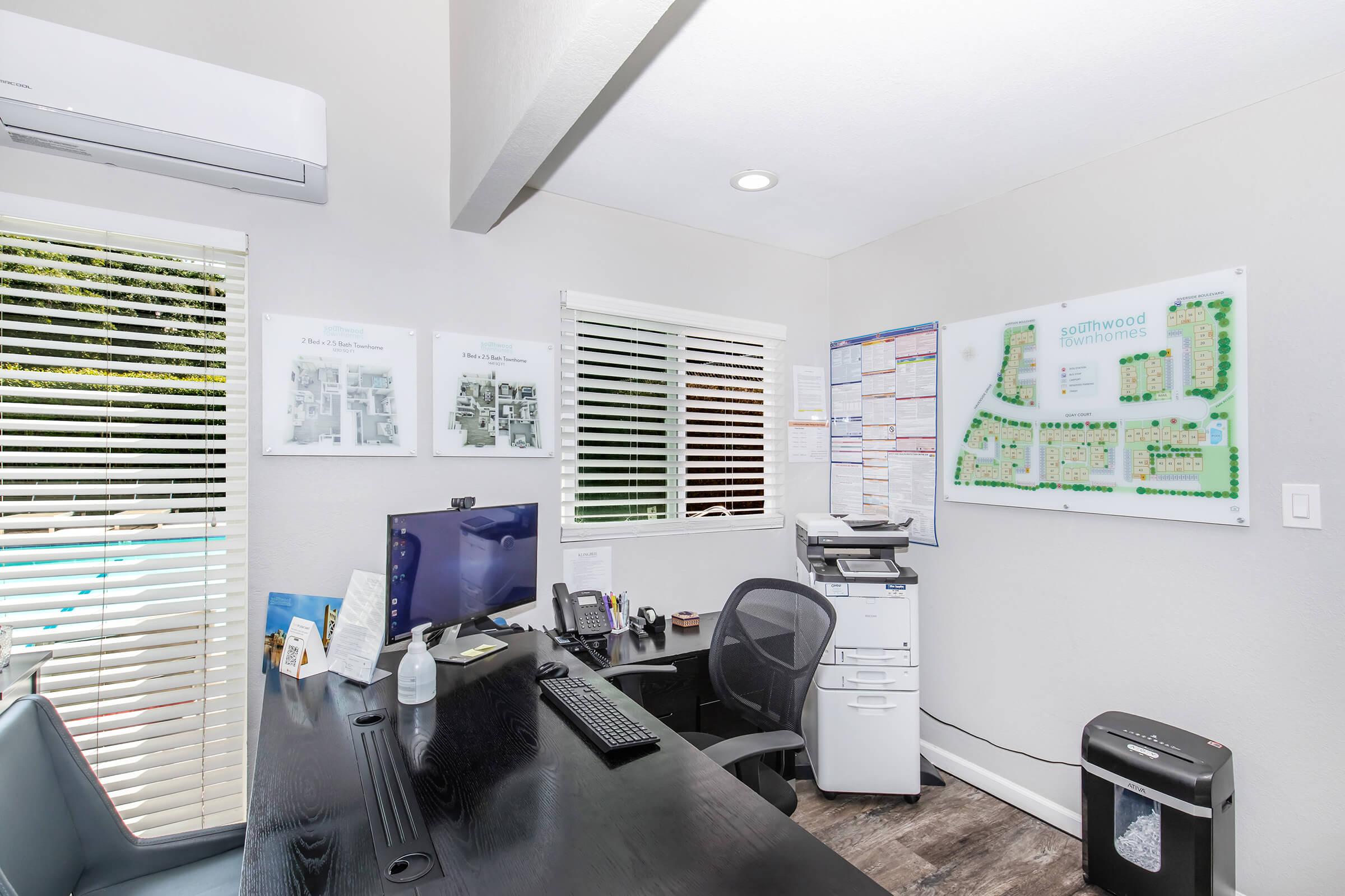 An office space featuring a black desk with a computer monitor, chair, and printer. The walls have posters and a large map on display. There are white blinds on the window allowing natural light, and a small shredder sits near the desk. The overall decor is modern and minimalistic.