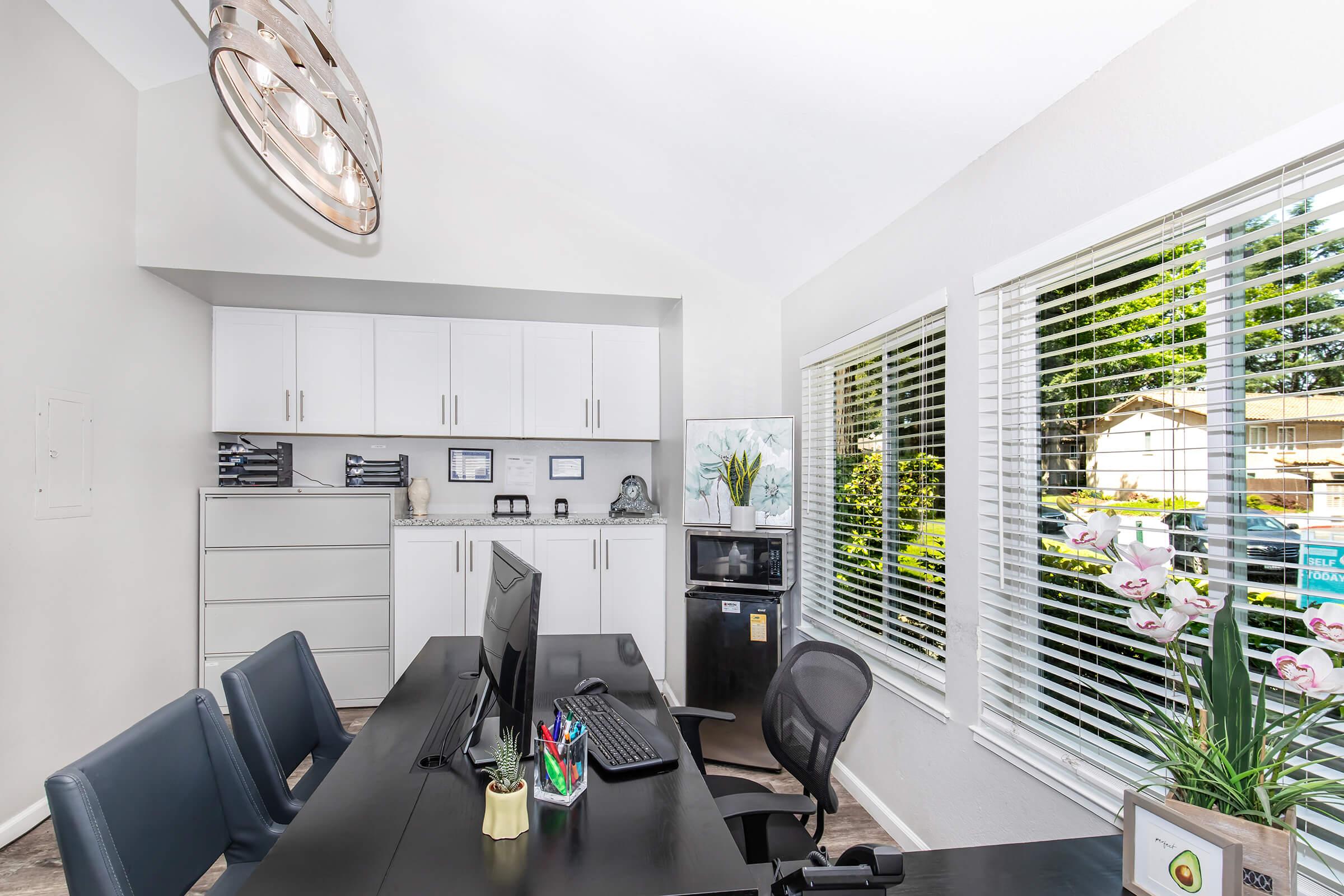 A modern office space featuring a black desk with a computer, organized stationery, and a chair. In the background, there's a water cooler, filing cabinets, and a large window with blinds that lets in natural light, revealing greenery outside. Decorative orchids add a touch of elegance to the desk area.