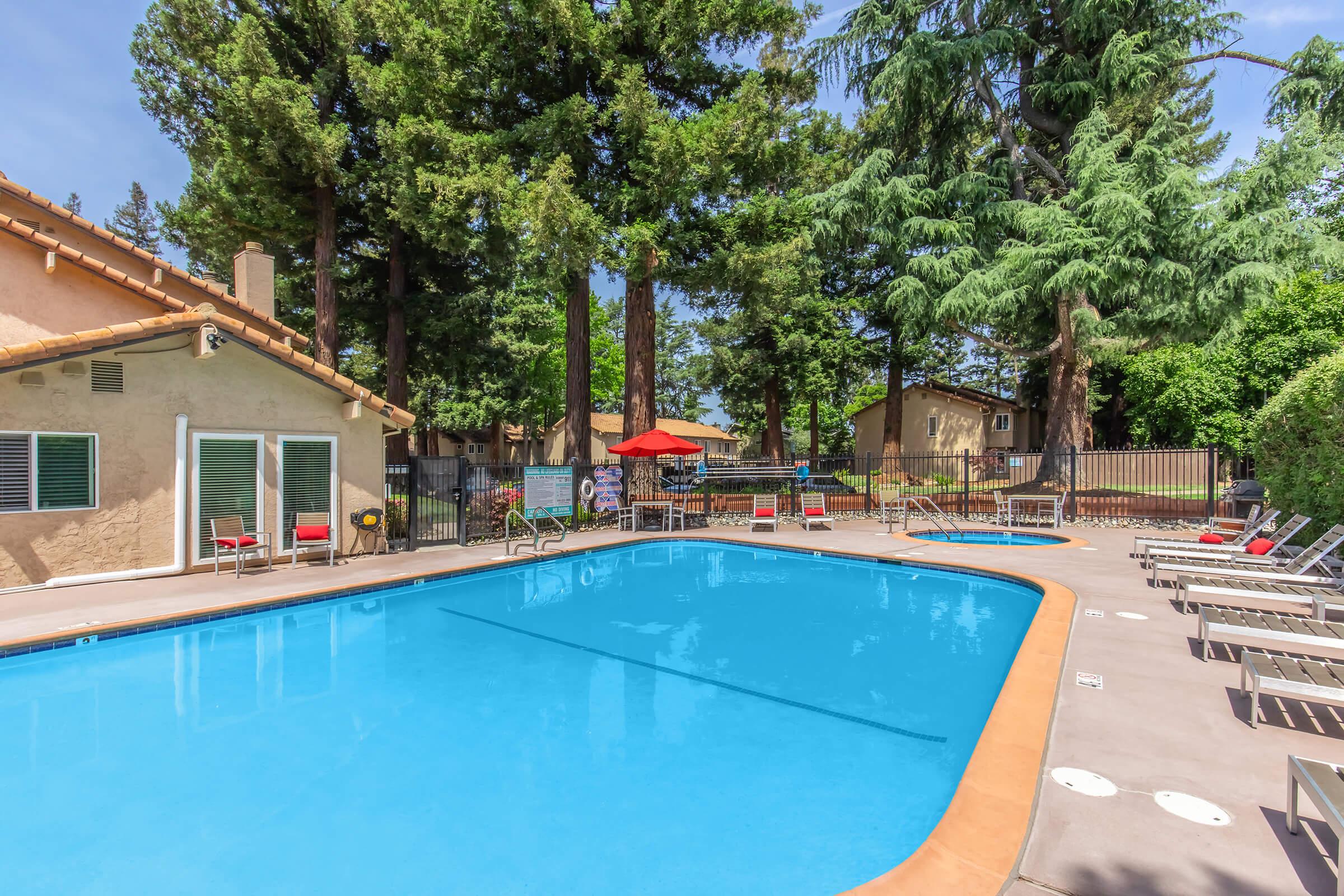 A clear blue swimming pool surrounded by tall trees and lounge chairs, with a few tables and umbrellas in the background. Several poolside chairs are neatly arranged. The area is well-maintained and inviting, ideal for relaxation or family gatherings. A fence encloses the pool area, enhancing privacy.