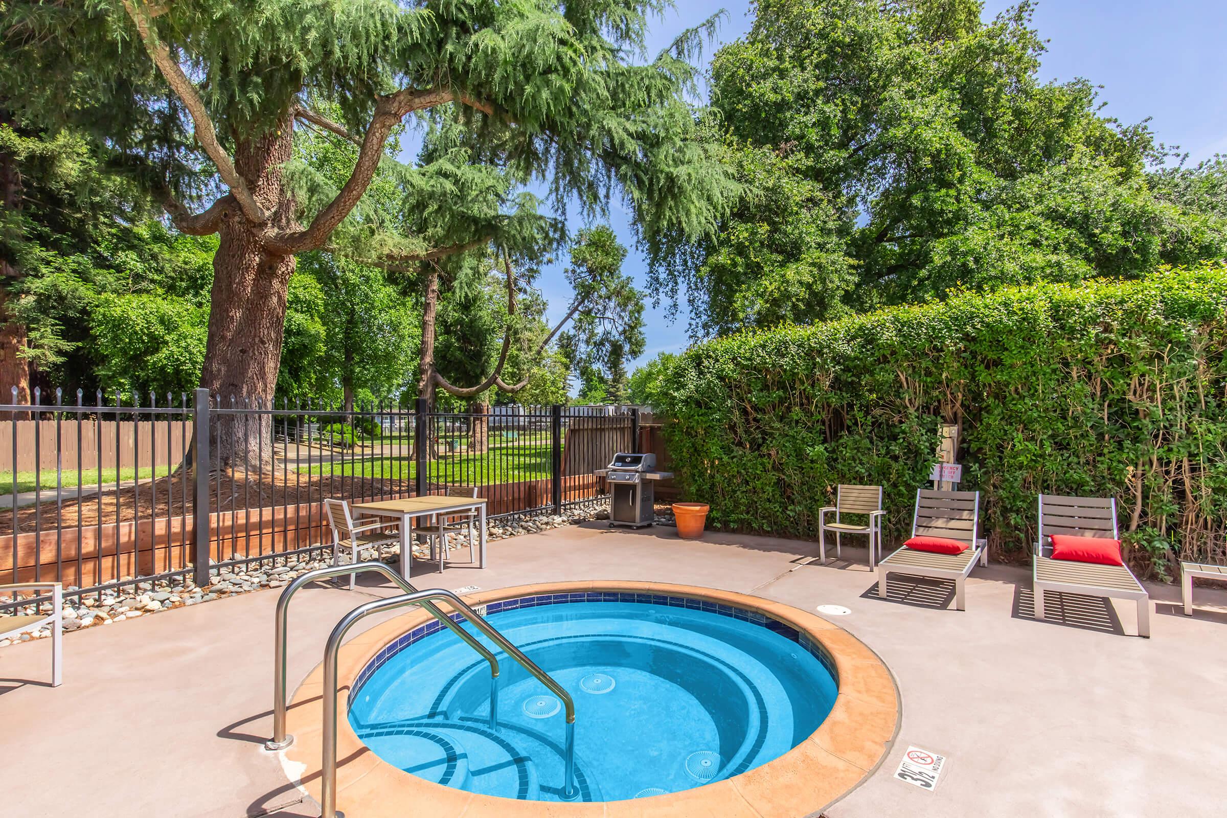 A serene outdoor spa area featuring a circular hot tub, surrounded by lush greenery and trees. Nearby, there are two lounge chairs with red cushions and a barbecue grill. The area is enclosed by a black fence, enhancing the privacy of this inviting relaxation space.