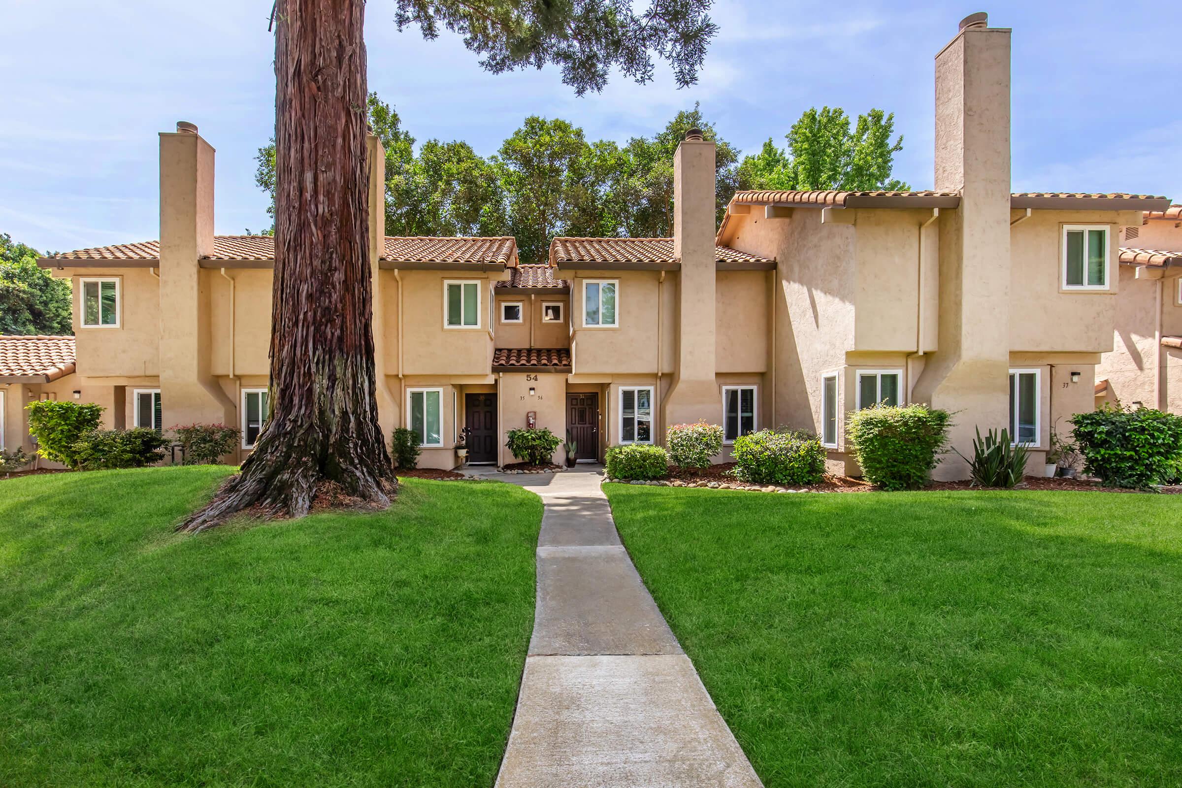 A residential complex featuring two-story townhouses with light-colored stucco exteriors, symmetrical windows, and prominent chimneys. A well-maintained green lawn and a pathway lead to the entrance, with trees providing shade in the background under a clear blue sky.