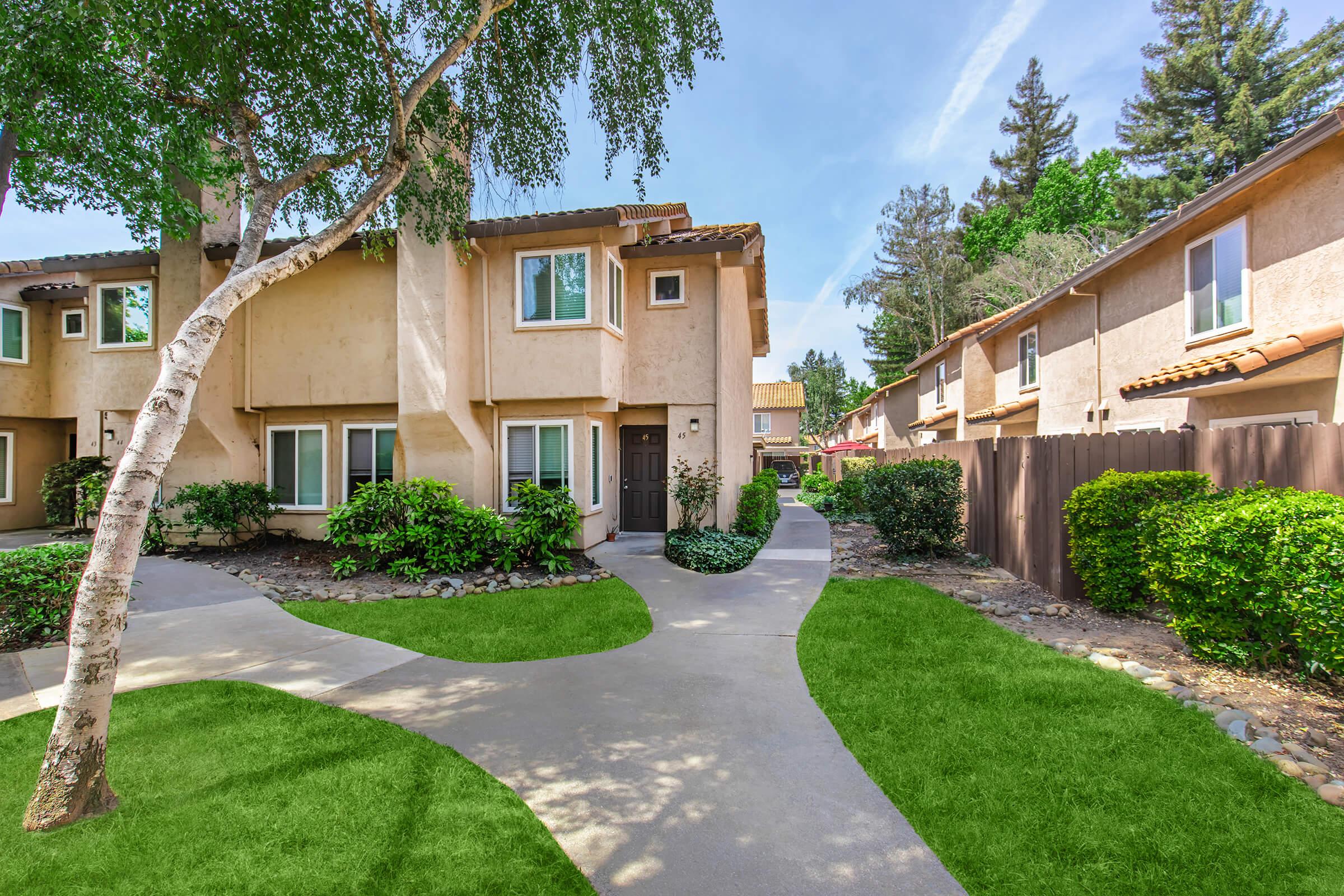 A landscaped pathway leading to residential buildings surrounded by green grass and trees. The area features well-maintained shrubs and a clear blue sky. The architectural style is modern with earthy tones, creating a welcoming environment.
